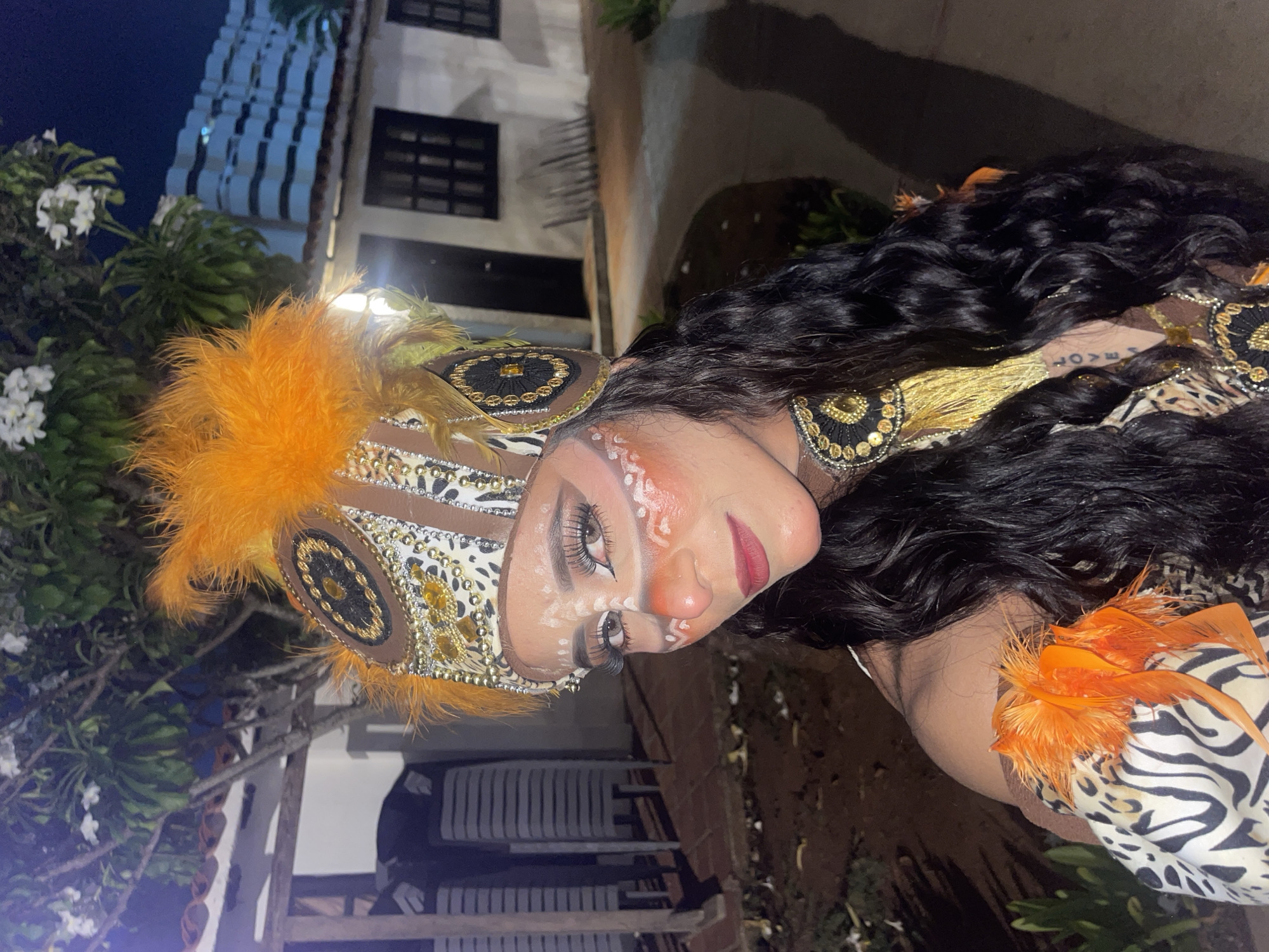 A woman, possibly of Venezuelan descent, is pictured outdoors at night in Porlamar, Venezuela. She is wearing a tribal-inspired costume and face paint. Her headdress is adorned with orange feathers and intricate beadwork. Her face is painted with white and orange markings, and she has dramatic eye makeup and dark lipstick. She has long, dark, wavy hair. Her costume features animal prints and gold embellishments, including a large, beaded necklace. In the background, a white building with dark windows and a tiled roof is visible, along with some trees and outdoor furniture. The overall scene suggests a festive occasion or performance.