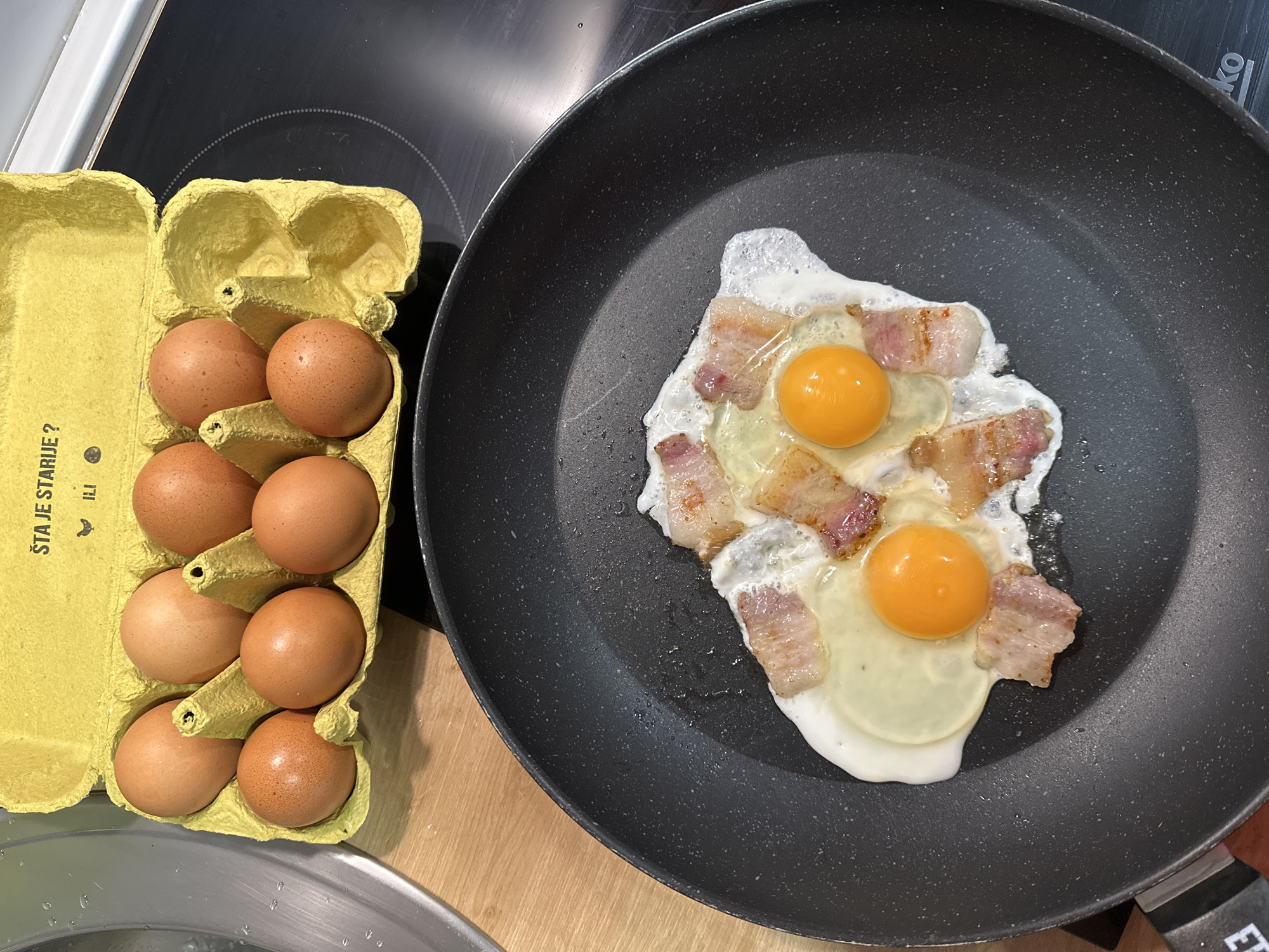 On a wooden surface, a black frying pan contains two fried eggs with bacon strips arranged around them. To the left of the frying pan is a yellow carton containing ten brown eggs. The scene is likely set indoors on a kitchen counter or stove top.