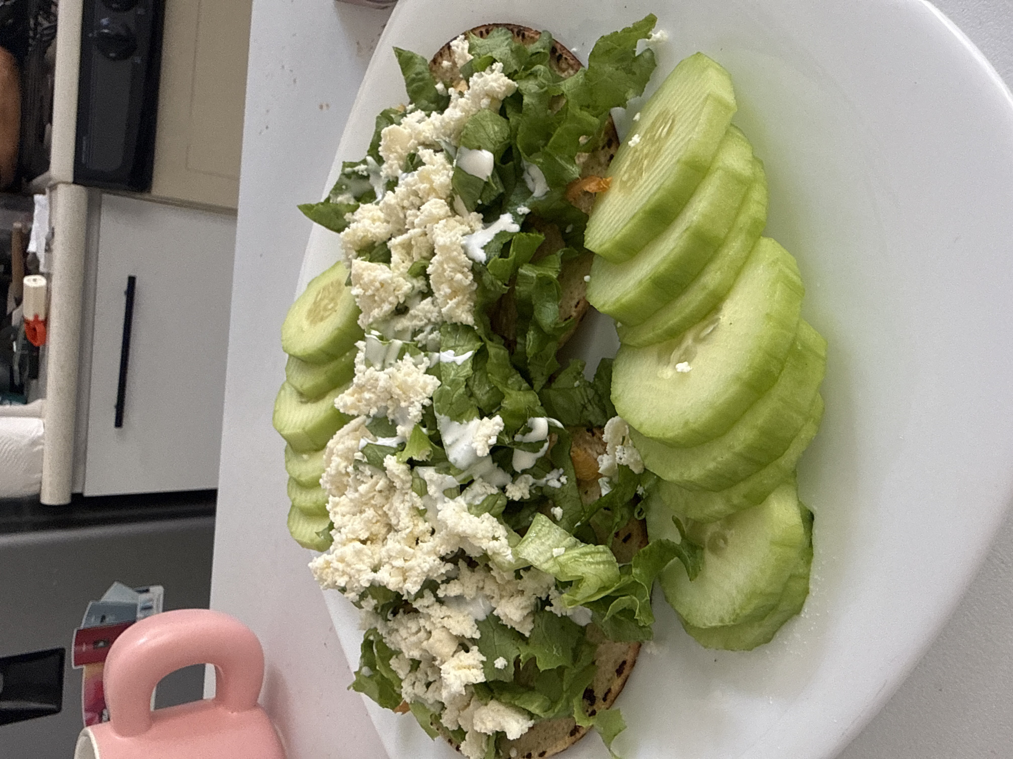 A close-up shot showcases a plate of food. The dish appears to be two tostadas. Each tostada is covered in lettuce, crumbled cheese, and a white sauce or cream. Sliced cucumbers are arranged along one side of the tostadas.
Without additional information, determining the exact location in Puebla, Mexico, is impossible. The environment in the image primarily consists of the food and the plate it is served on. The background is blurred, showing a white refrigerator and a pink mug on the counter.