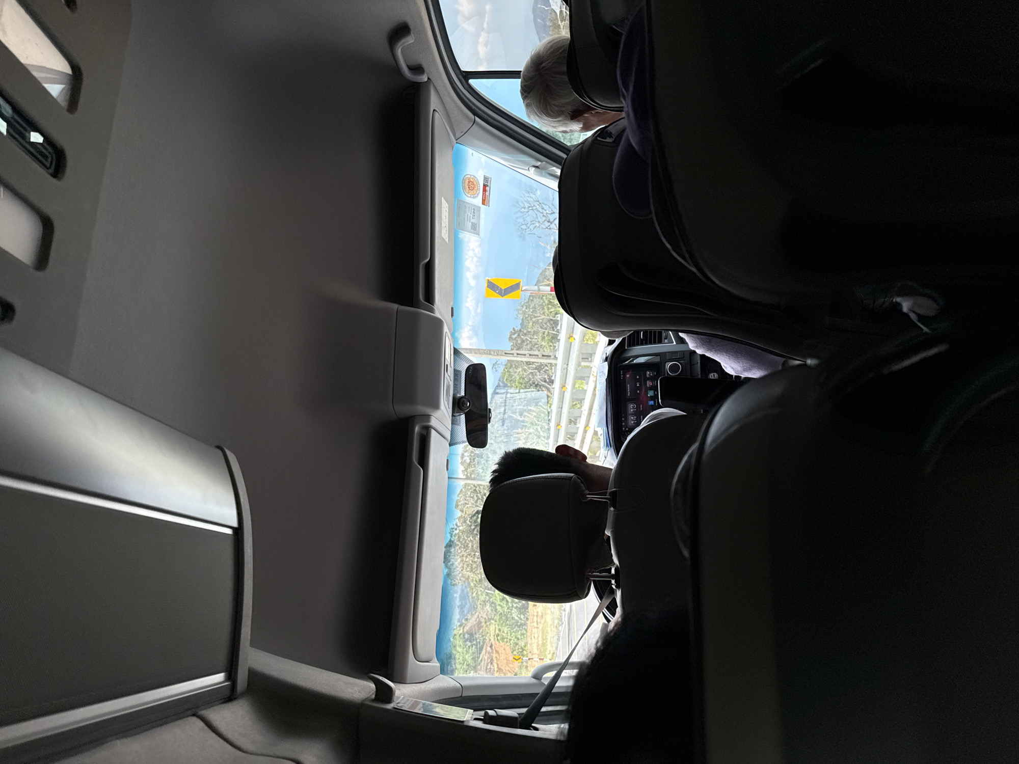 The image depicts the interior of a vehicle traveling on a road in or near Nam Khánh Vĩnh, Vietnam. In the foreground, the backs of several seats are visible, some with passengers. The central focus is on the driver, a male with dark hair, wearing a light-colored shirt, and the headrest of his seat. The dashboard of the vehicle is also visible, displaying a clock at 14:13. Through the windshield, a winding road with yellow directional signs is seen, bordered by a guardrail and lush green hills with scattered trees. The sky is partly cloudy, suggesting daytime travel. To the right, the head and shoulder of a passenger with gray hair are visible, looking out the window. The interior of the vehicle appears to be a van or similar passenger transport.