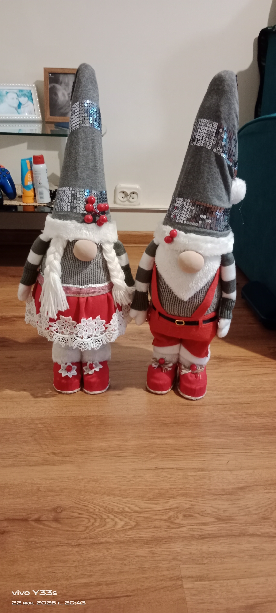 This media file features two decorative gnomes, likely intended for holiday display, standing side-by-side on a wooden floor. The gnome on the left is dressed in a gray conical hat adorned with sequins and red berries, a white fuzzy trim, and striped sleeves. It wears a gray knitted dress with a red skirt trimmed with white lace and red boots with white flower details. White braided pigtails hang from its sides. The gnome on the right wears a similar gray sequined hat with a white pom-pom, white fuzzy trim, and striped sleeves. It is dressed in a gray patterned vest over a white shirt, red overalls with a black belt, and red boots with red embellishments. Both gnomes have round, light-colored noses and appear to be holding hands.
The background is a plain, light-colored wall with an electrical outlet visible. To the left, on a shelf, are some framed pictures and a few bottles, including one that appears to be sunscreen. The overall impression is of a domestic setting, possibly a home interior.
The lighting suggests it could be daytime, as the scene is well-lit, but there are no direct indicators of natural light or weather conditions. The presence of the gnomes suggests a festive or seasonal context, likely related to winter holidays.
Visible text at the bottom of the image reads "vivo Y33s" and "22 июн. 2026 г., 20:43". This indicates the device used to capture the image and a date and time stamp, suggesting the photo was taken on June 22, 2026, at 8:43 PM.
The location context provided is Oława, Poland.