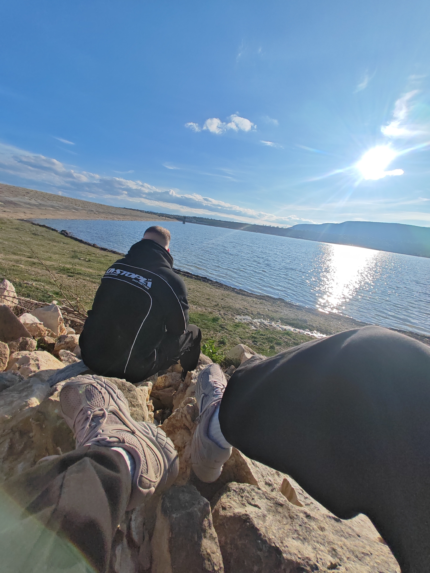 A wide-angle shot captures two individuals by a lake on a sunny day. The viewer's perspective is from one person's point of view, with their legs and feet visible in the foreground. They wear grey trainers and black trousers, resting on rocks. Beside them, another individual sits with their back turned, wearing a black hoodie with white detailing on the back. The person is positioned closer to the lake.
The lake extends into the distance, reflecting the sunlight and creating a sparkling effect on the water's surface. In the background, a hilly landscape and mountains are visible across the lake. The sky is blue with scattered clouds. The foreground consists of rocky terrain and patches of grass near the lakeshore.
The location is likely near a dam in Shumen, Bulgaria.
