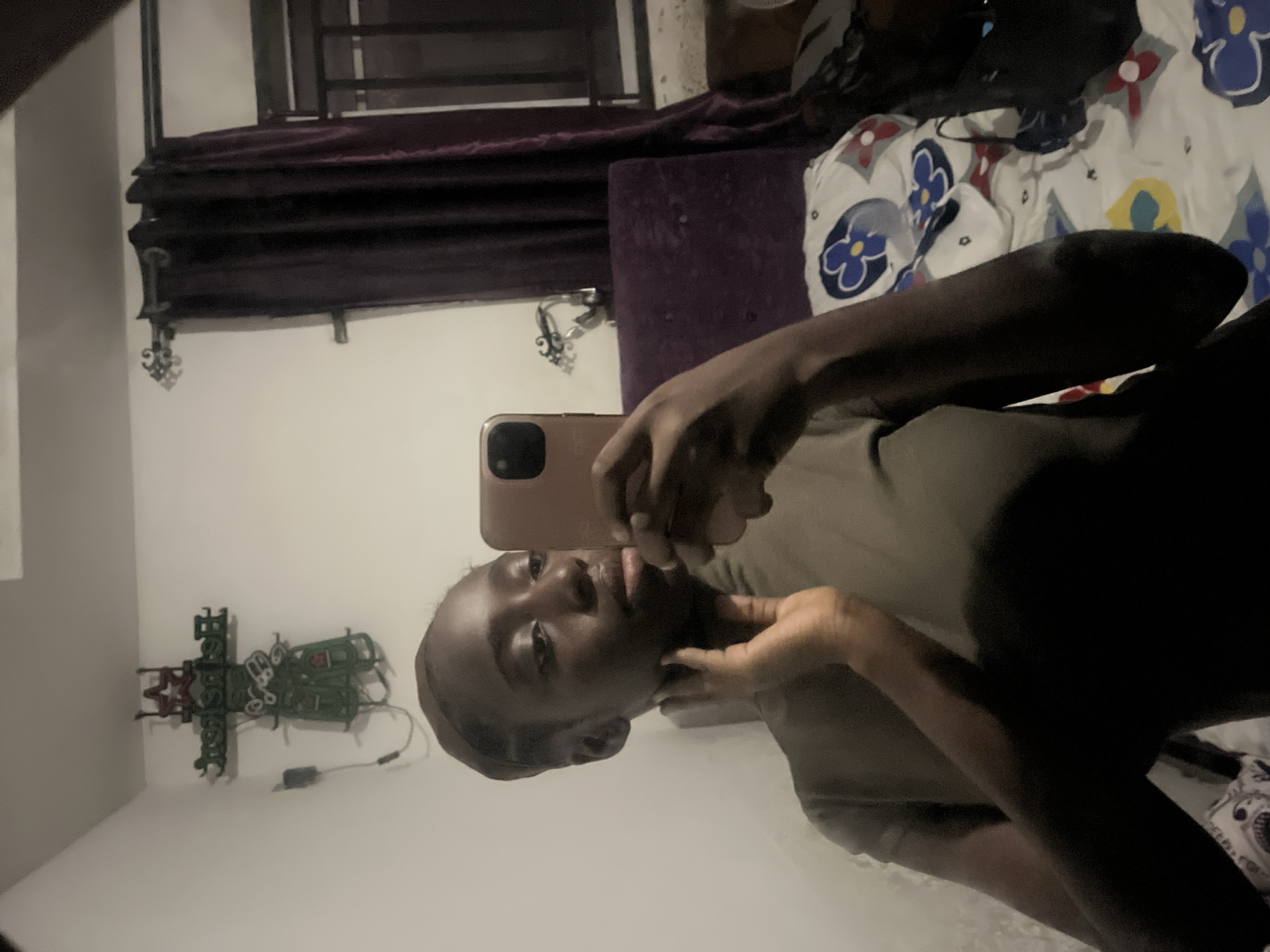 A young, dark-skinned person is taking a selfie in what appears to be a bedroom. The person is holding a beige cell phone and posing with their hand near their face. The room contains a bed with patterned white, blue, and yellow sheets, a purple headboard, dark purple curtains, and an ornamental wall hanging of a beer sign. Given the image's origin, it's plausible the location is in Abuja, Nigeria.