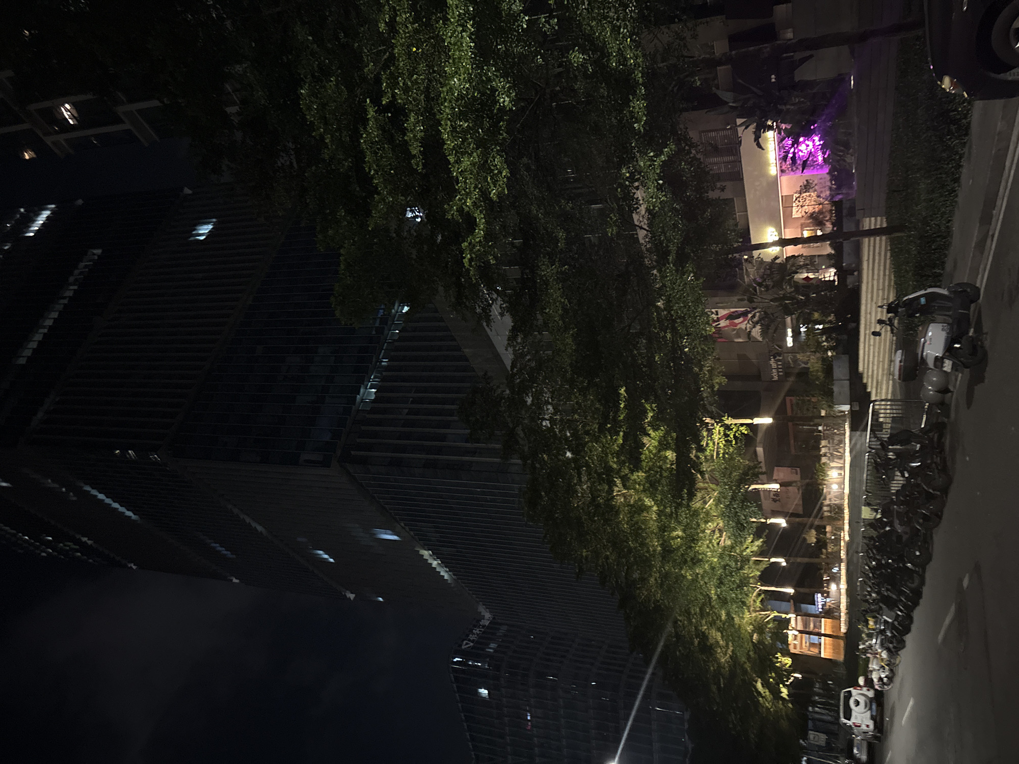 The media file captures a nighttime scene in Haizhu District, China. The image is oriented vertically, showcasing a cityscape with a prominent modern building characterized by its dark, horizontal slatted facade. Several illuminated windows are visible on the building, hinting at activity within.
Adjacent to the building is a dense canopy of lush green trees, their foliage casting shadows and adding a natural element to the urban environment. A street runs alongside the trees, lined with parked motorcycles and a white SUV. The street is illuminated by various light sources, including streetlights, building lights, and a vibrant purple light emanating from a structure visible in the distance. This purple light suggests a commercial establishment or entertainment venue.
The time of day is clearly night, indicated by the dark sky and artificial lighting. The weather appears to be clear, with no visible signs of rain or fog. The scene evokes a sense of urban vibrancy and nighttime activity, with the contrast between the dark, imposing building and the illuminated trees and street creating a dynamic visual.
There are no clearly discernible people in the image, though the presence of vehicles suggests human activity. No specific event is depicted, but the overall impression is one of a typical evening in a developed urban area.
Visible text is minimal and mostly illegible due to the image quality and distance. However, a few signs are partially visible on the buildings across the street, though their content cannot be determined. The overall atmosphere is one of a bustling city at night, with a mix of modern architecture and natural elements.