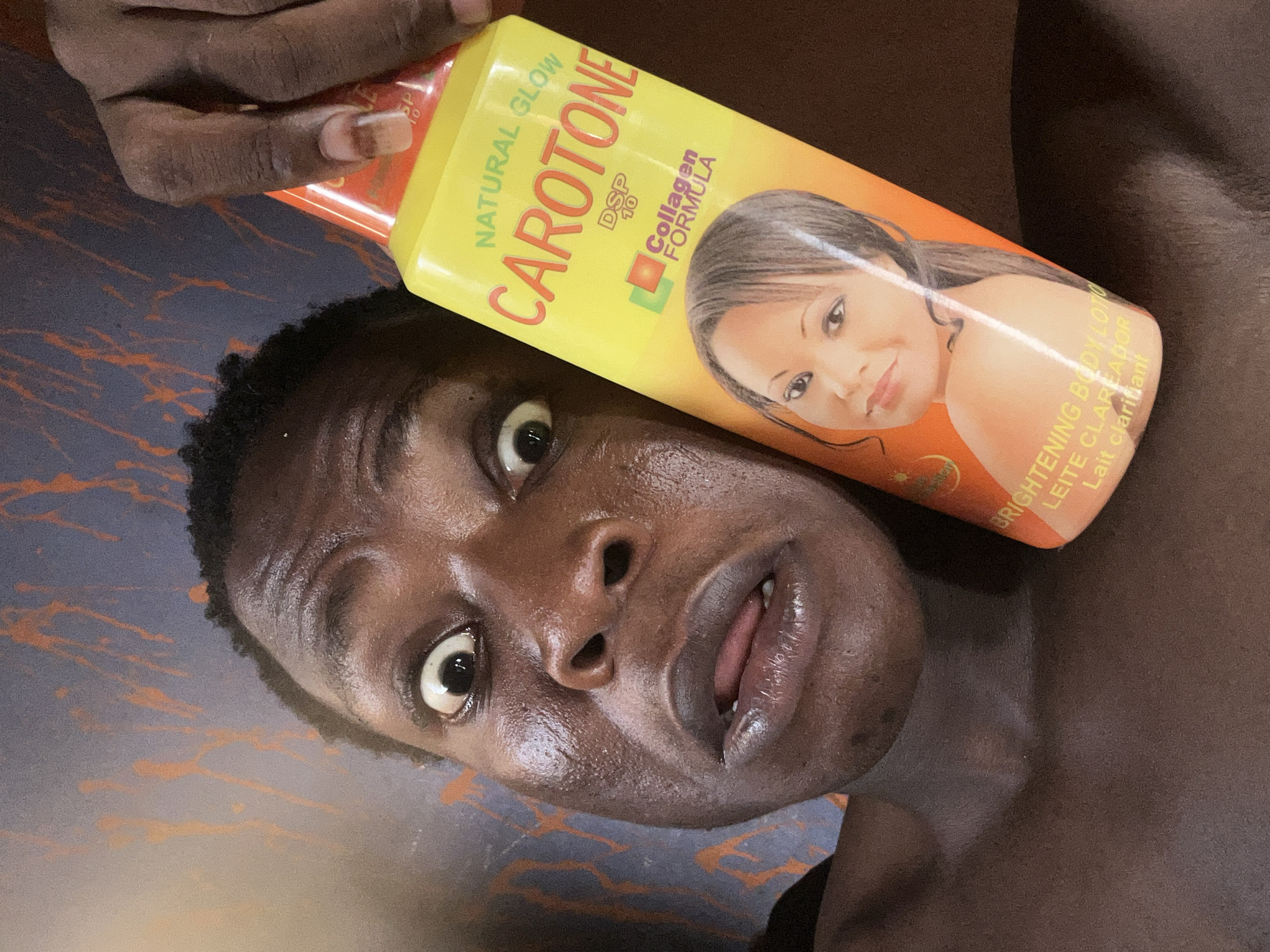 A close-up shot from a low angle captures a person holding a yellow bottle of "CAROTONE" Natural Glow DSP 10 Collagen Formula skin-lightening lotion. The person's face is glistening with what appears to be sweat or lotion. The person's expression includes wide eyes and an open mouth. The bottle is tilted towards the camera, partially obscuring the background. The background is a dark surface with abstract orange and blue patterns. The specific location and city are unknown.