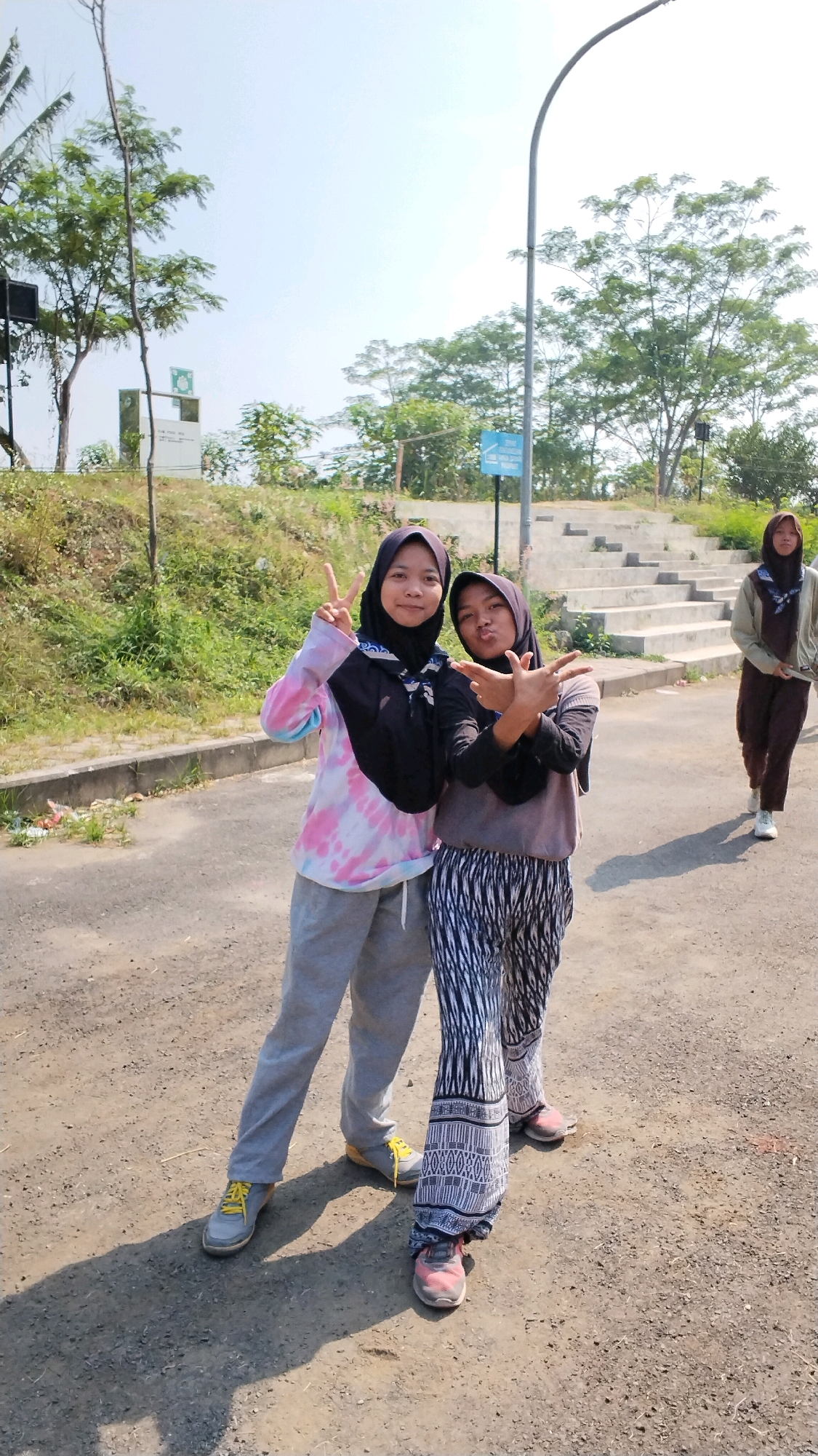 Two young women wearing hijabs pose for a photo in an outdoor urban setting. The woman on the left wears a tie-dye t-shirt with pink and blue colors and grey sweatpants. She is making a peace sign with her right hand. The woman on the right wears a grey long-sleeved shirt and patterned pants. She is making a gesture with both hands in front of her. They are standing on a dirt and asphalt path. In the background, there are stairs, trees, a streetlamp, and a sign. Another woman wearing a hijab and a light brown jacket is walking away from the camera in the background. The location is identified as Ngingasrembyong, Indonesia. The sky is clear and blue, suggesting it is daytime.