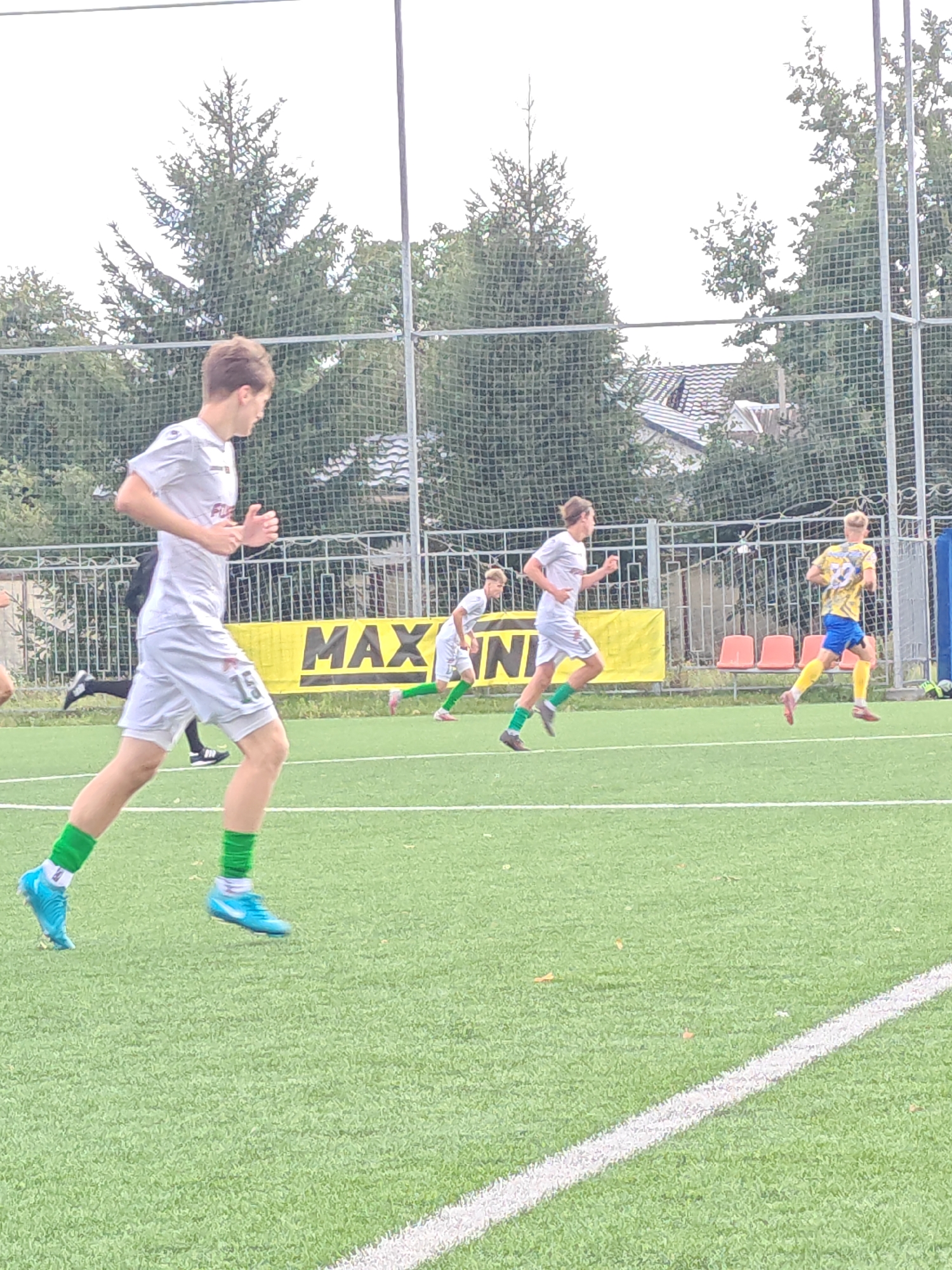 In Homyel, Belarus, this image captures a soccer match on an artificial turf field. In the foreground, a young male athlete in a white jersey with the number 15 visible on his shorts and green socks, runs towards the left of the frame. He is wearing blue cleats. To his right, another young male athlete in a yellow and blue jersey with green socks and pink cleats runs towards the right. In the background, a yellow banner with the text "MAX N" is visible. Trees and a chain-link fence border the field. The sky is overcast.