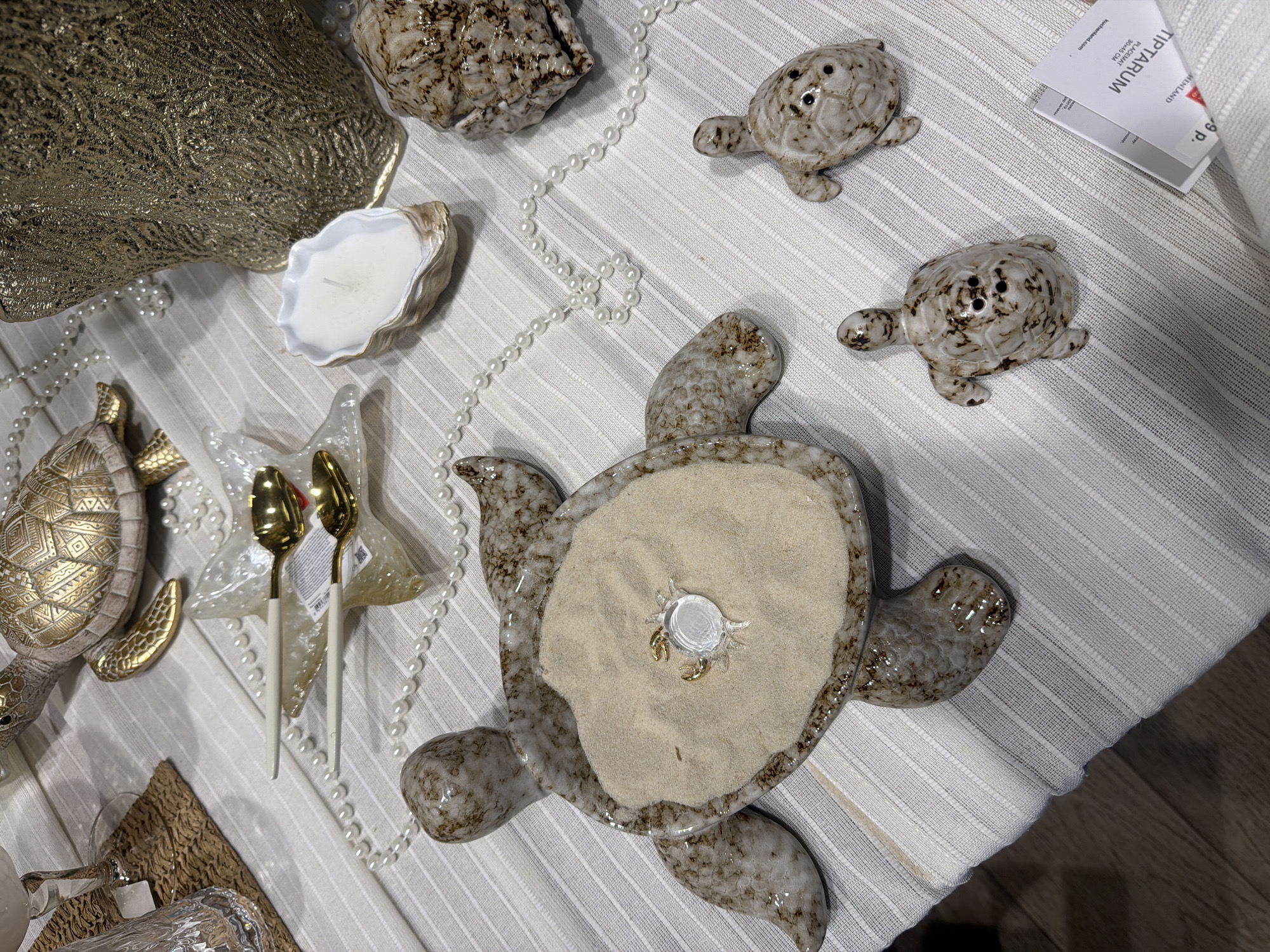 The image presents a still life arrangement on a white, horizontally striped surface. Several decorative elements are scattered across the scene. There's a large, textured golden object, possibly a platter, occupying the upper left corner. To the right of it, a beige-and-white shell-shaped object, likely a candle holder with a white candle inside, is visible. A string of white pearls meanders around these elements. Two small turtle figurines, with mottled brown patterns, are situated near the top of the frame. Further down, a larger, sand-filled turtle dish with a small, clear plastic crab toy in the center is placed. Two golden spoons with white handles lie beside a clear, starfish-shaped dish. To the lower left, there's a partially visible decorative turtle, adorned with gold patterns. An additional object made of woven material and a clear glass container with a metallic cap are situated at the very bottom left corner. A small white card with the text "TIPTARUM" is placed next to one of the turtle figurines. The setting suggests a decorative display. The mention of Novoalexandrovka, Russia is irrelevant, as the scene is contained within a still life arrangement and does not provide geographic context.