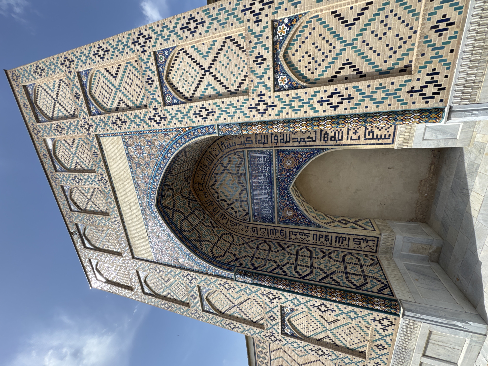 The image shows the facade of a building with intricate tilework in Samarkand City, Uzbekistan. The structure is decorated with a pattern of small rectangular tiles in shades of beige, blue, and black. There is a large arched entrance adorned with floral motifs and Arabic script. The sky is visible in the background, with a few light clouds. The architectural style appears to be Islamic, likely part of a historic monument or religious building.