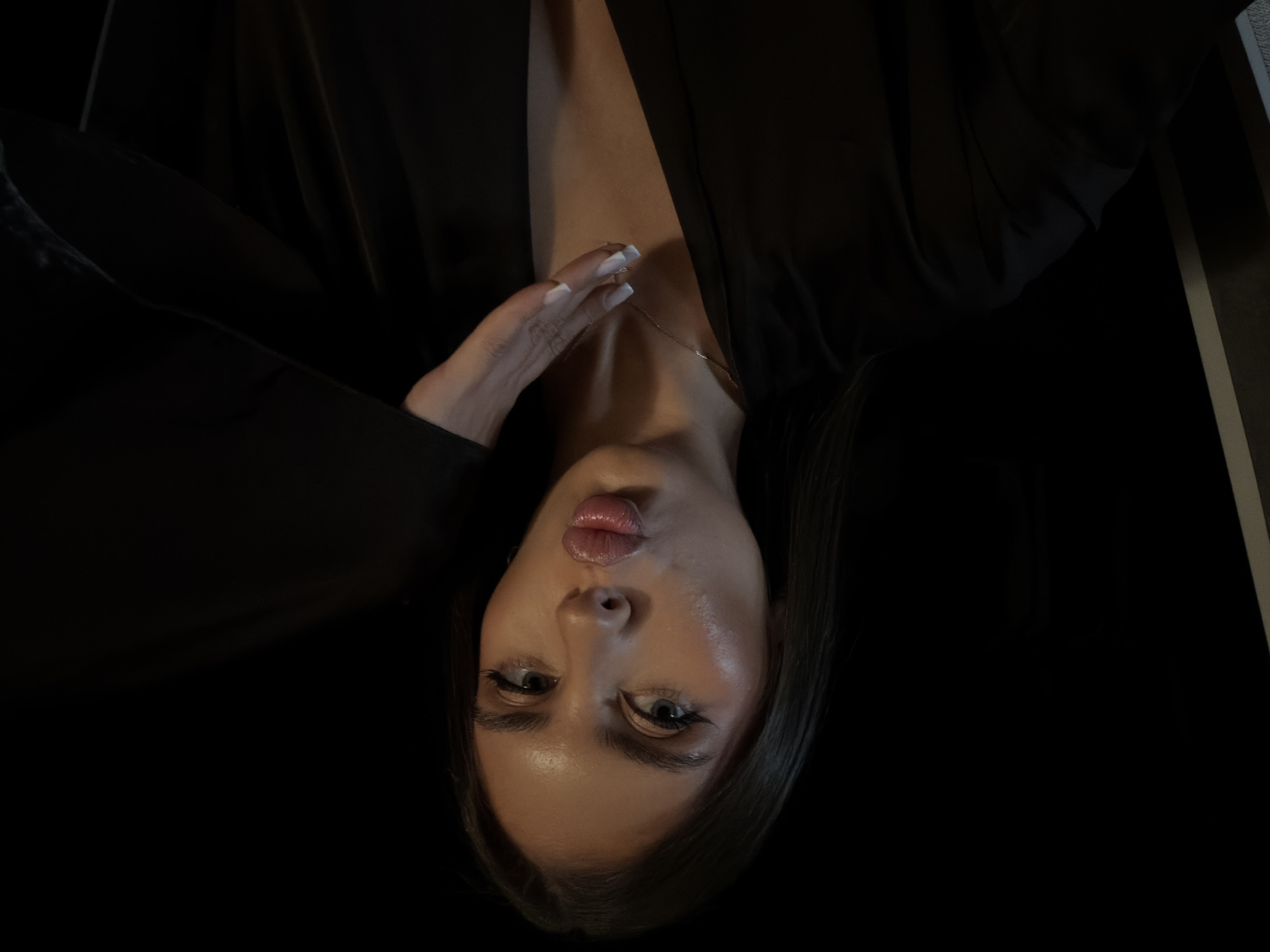 A woman with fair skin, dark hair, and a small gold necklace is positioned at a rotated angle in the image. She has full lips pursed in a kissing expression, and her eyes are open, looking directly at the viewer. Her left hand is raised near her neck, with her fingers extended. She is wearing a dark-colored jacket or blouse with a deep V-neck. The background is dark, suggesting a dimly lit indoor setting. There is no visible evidence of Krasnoyarsk, Russia.