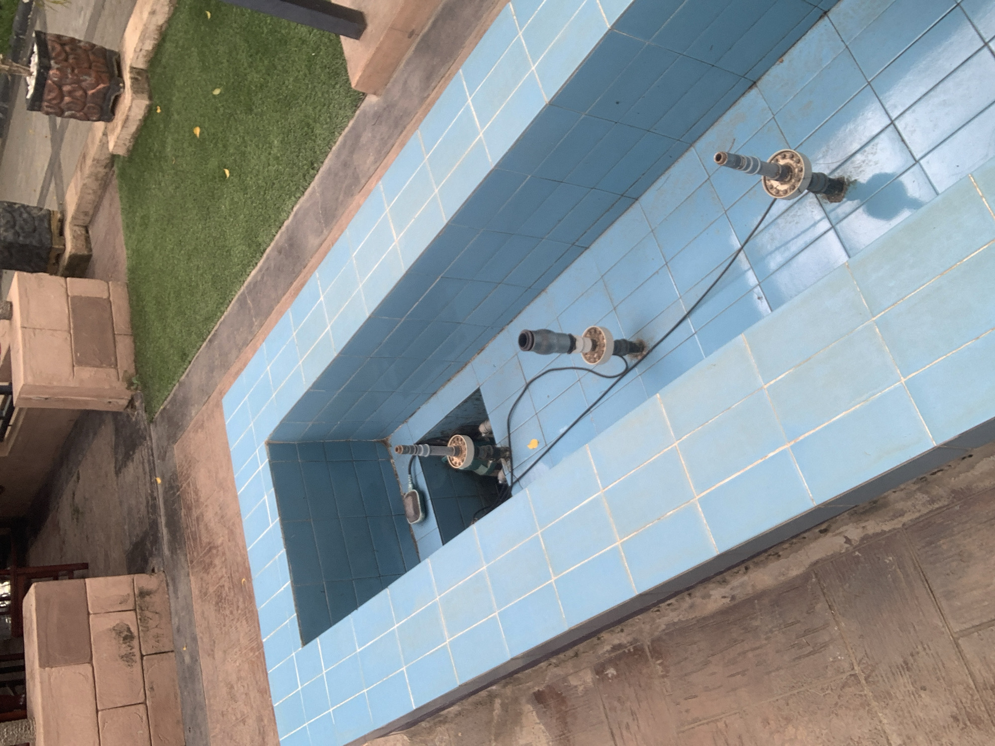 The image shows a dry, tiled fountain structure in an outdoor setting, possibly a public space or a residential compound, in Abuja, Nigeria. The fountain basin is rectangular with light blue tiles. Inside the basin, there are several spouts or nozzles, some appearing to be connected to plumbing or electrical components. In the background, there is an area of artificial turf, a stone retaining wall, and brickwork. The ground surrounding the fountain is paved with reddish-brown interlocking tiles. The scene is devoid of water, indicating the fountain is not operational at the time of the photograph. The overall impression is one of disuse or maintenance.