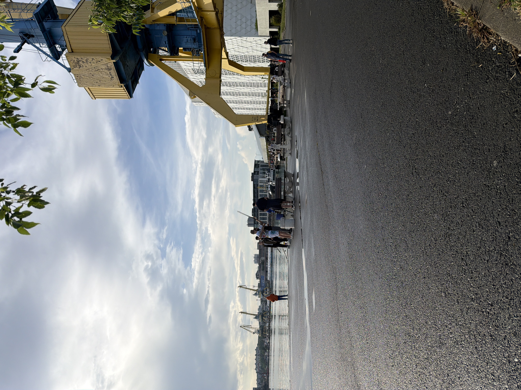 The image depicts a street scene in Burgas, Bulgaria, likely near the port area. In the background, several harbor cranes are visible along the water's edge, against a backdrop of city buildings. In the foreground, a group of people stand near the water, with some appearing to be fishing. A large yellow industrial crane looms over the scene on the left side of the frame. The ground is paved and appears damp, possibly from recent rain. The sky is partly cloudy, with blue sky visible behind the clouds.