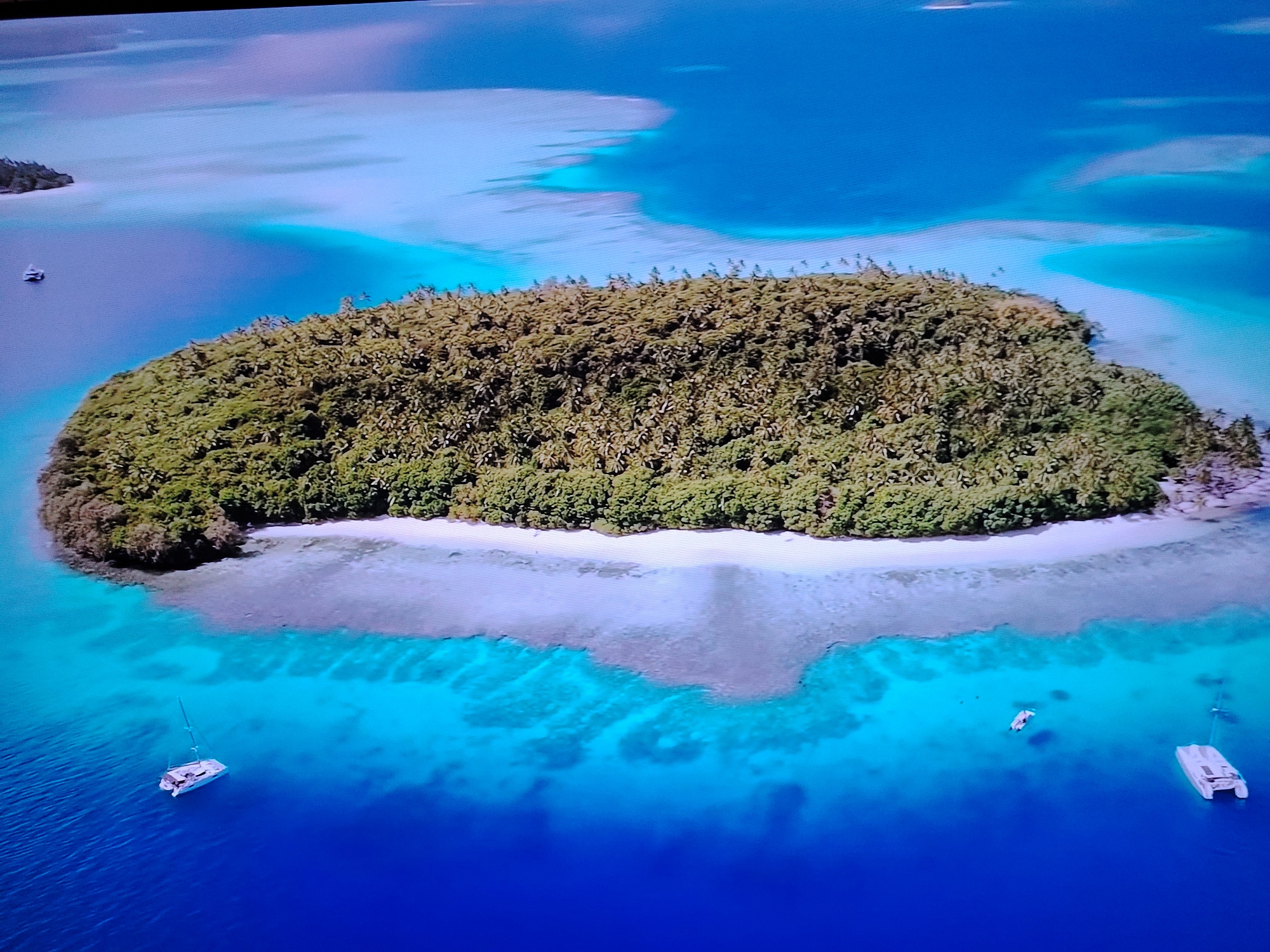 This is an aerial drone shot of a tropical island in a clear blue ocean. The island is densely covered with green foliage, likely palm trees. A white sandy beach lines a portion of the island. The water transitions from light turquoise near the shore to a deep blue further out. There are three boats visible in the water: a sailboat with a white hull and mast in the lower left, a smaller white boat in the middle, and a white catamaran on the right. In the background, other small islands and sandbars are visible in the turquoise water. The scene is devoid of any structures or people on the island or in the immediate vicinity. There is no indication that this scene is located in Huntingdonshire, United Kingdom.
