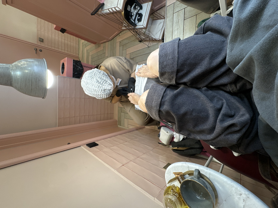 This image, captured from a first-person perspective, depicts a pedicure service underway within a beauty salon. The individual's bare feet, positioned in the foreground, rest on a white cushion or towel, with dark grey sweatpants rolled up to just below the knees. A dark grey fabric partially covers the individual's lap.
A technician, seated on a low stool facing the feet, is actively performing the pedicure. This individual wears a light brown uniform, a white disposable hairnet, and black gloves, with a face mask visible below their chin.
The salon interior features light pink-beige painted walls, with a lower section covered in horizontal light pink tiles. The floor is tiled in a light-colored herringbone pattern. An industrial-style grey metal pendant lamp hangs directly overhead, illuminating the workspace. To the left, a small white round table holds a clear glass jar containing a yellow liquid, a grey ceramic cup on a wooden coaster, and a small dark rectangular object. Below the table, a white electric nail drill or similar device is visible. In the background, a light pink cylindrical trash can with a black liner is present, bearing partial Russian text, "ОГНЕМИНА" (OGNEMINA), identifying the location as a salon belonging to this chain in Moscow, Russia. Further right, a tall pink cabinet or display unit stands, and a light pink door with a silver handle is visible in the background.