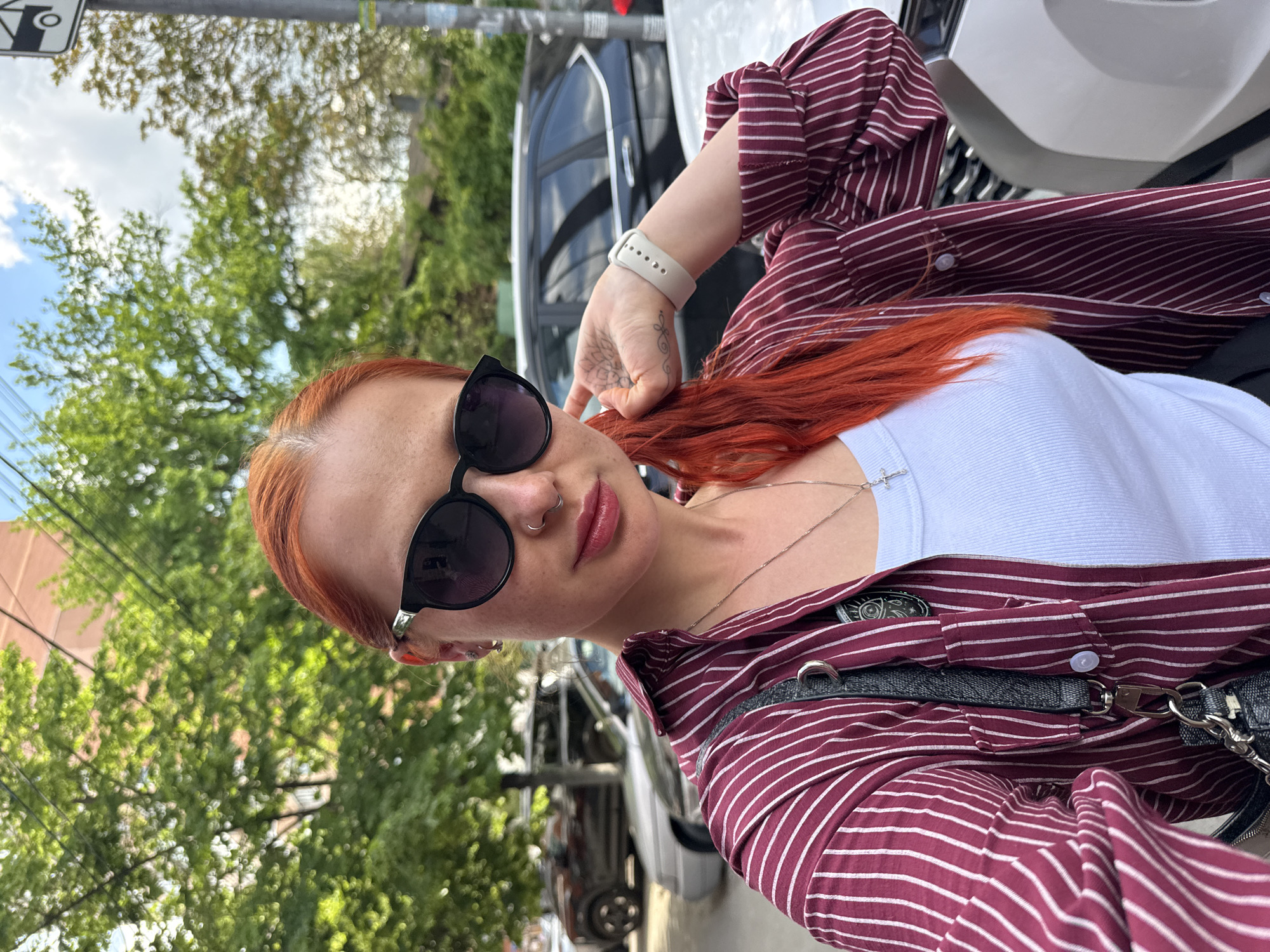 A woman with orange hair stands on a city street. She wears a white tank top under a striped maroon shirt. Her hand rests on her hair, and she wears sunglasses and a silver necklace with a cross pendant. The street is lined with trees and parked cars. The city is Rostov-on-Don, Russia.
