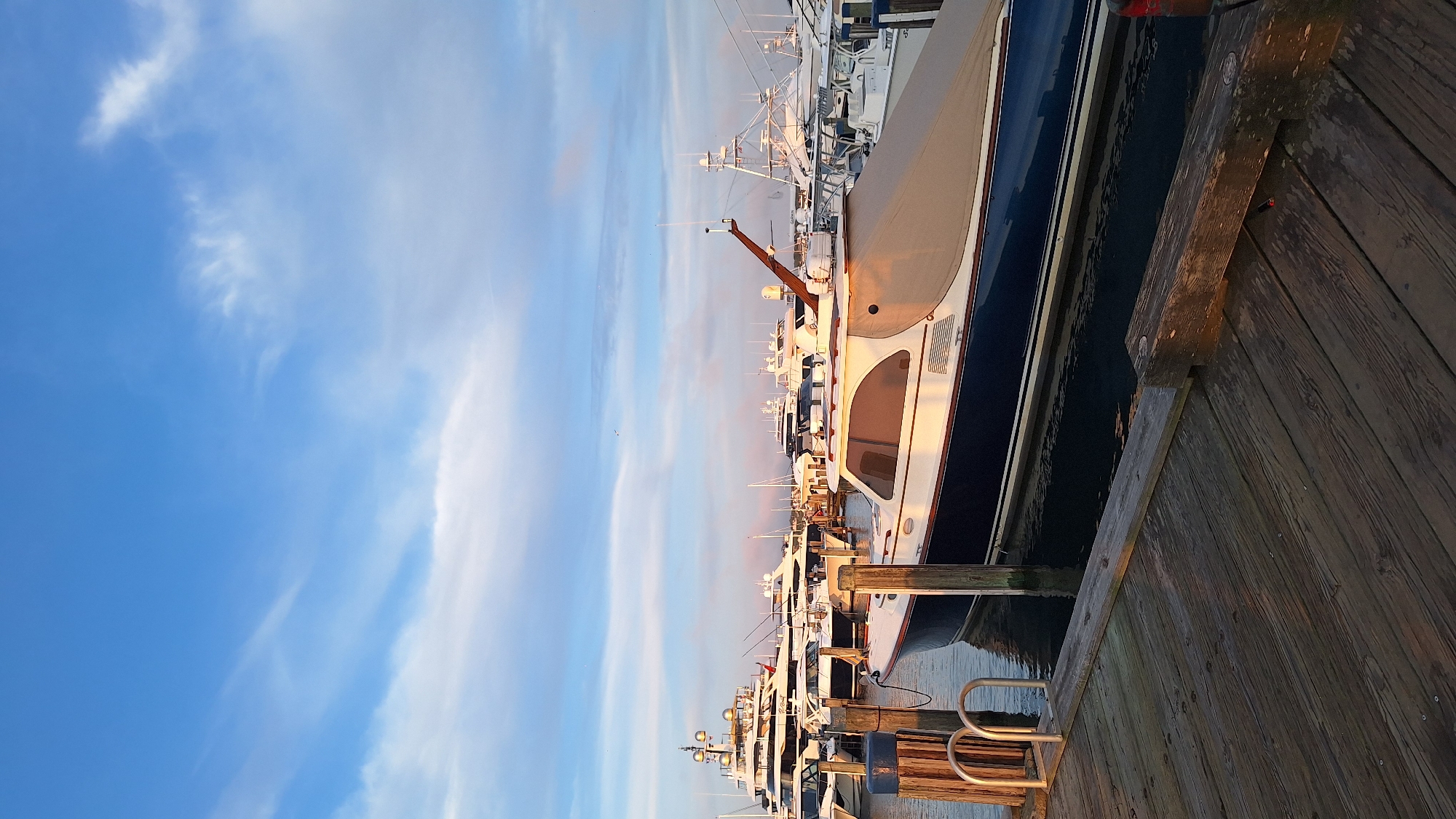 The image shows a marina on Nantucket, a small island located off the coast of Massachusetts, USA. The scene is dominated by several large yachts and smaller boats docked in the water. In the foreground, a wooden dock is visible, with a portion of a blue and white boat tied up to it. The boat is covered with a tan tarp, and a small metal ladder is attached to the dock. In the background, a large white yacht with multiple levels and a prominent radar array is docked. The sky is blue with scattered white clouds, and the lighting suggests it is either early morning or late afternoon, with the sun casting a warm glow on the scene. The overall atmosphere is peaceful and serene, characteristic of a quiet harbor.