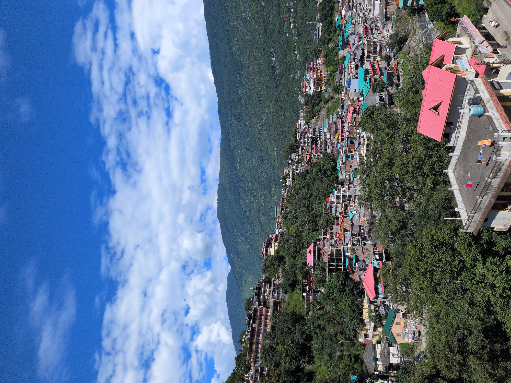 The image shows a densely populated hillside town in Kullu, India, with buildings cascading down the slope. The architecture is a mix of traditional and modern styles, with many houses featuring colorful roofs, predominantly red and green. The town is nestled amidst lush green mountains under a bright blue sky dotted with white clouds. Numerous trees are interspersed throughout the town and on the surrounding hillsides. In the foreground, a house with a prominent red roof is visible, with laundry drying on its rooftop. Vehicles are present on the roads, and some people can be seen, though they are not the primary focus of the image. The overall scene conveys a sense of vibrant life within a scenic, mountainous environment.
