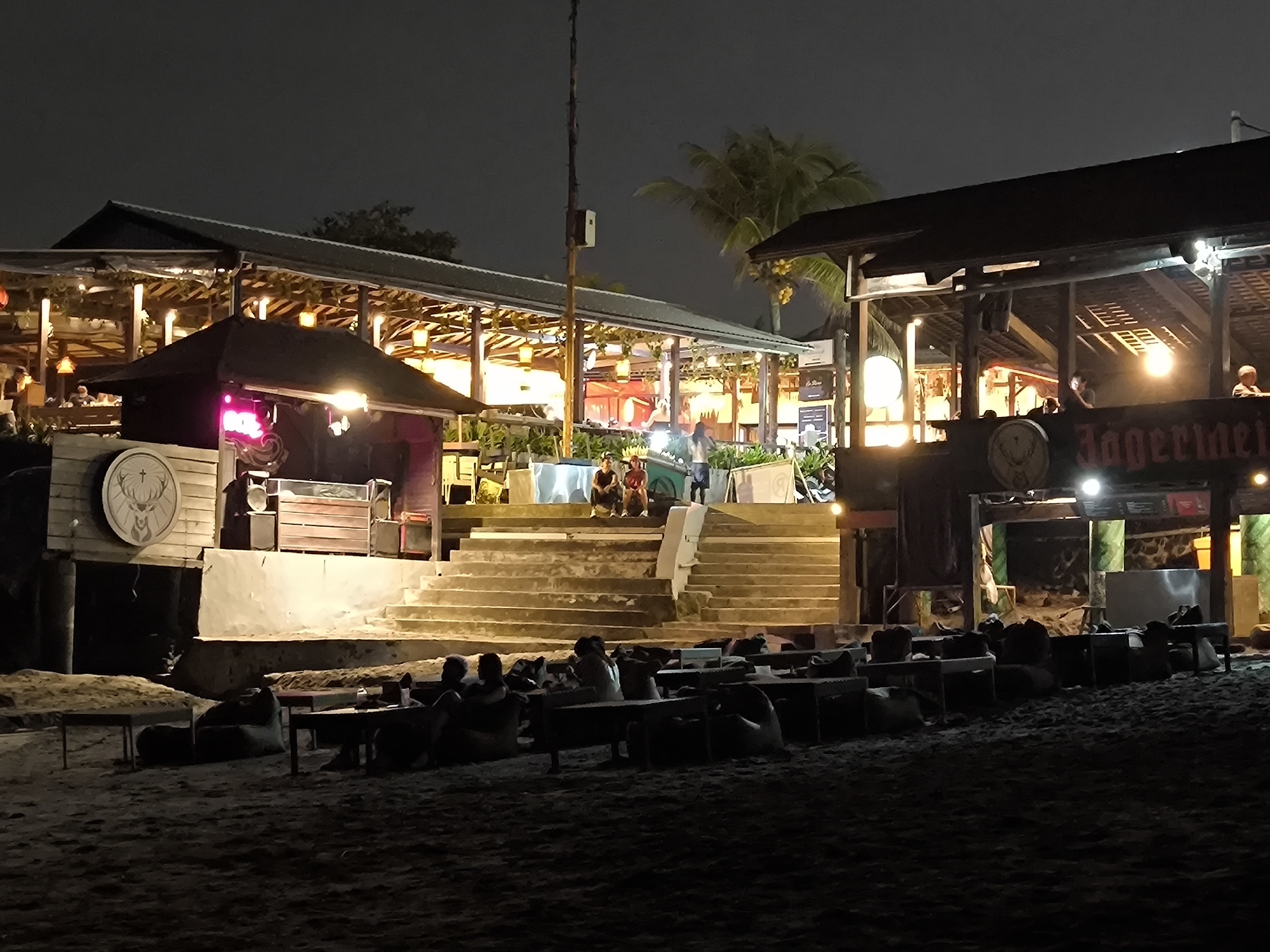 The image depicts a beachfront bar or restaurant in Canggu, Indonesia, at night. Several open-air structures with illuminated interiors are visible, one featuring a prominent Jägermeister logo. Groups of people are seated at low tables on the sand, some using beanbag chairs. A set of stairs leads up to the main area of the establishment, where more people are gathered. The overall atmosphere is relaxed and social. Palm trees are visible in the background.