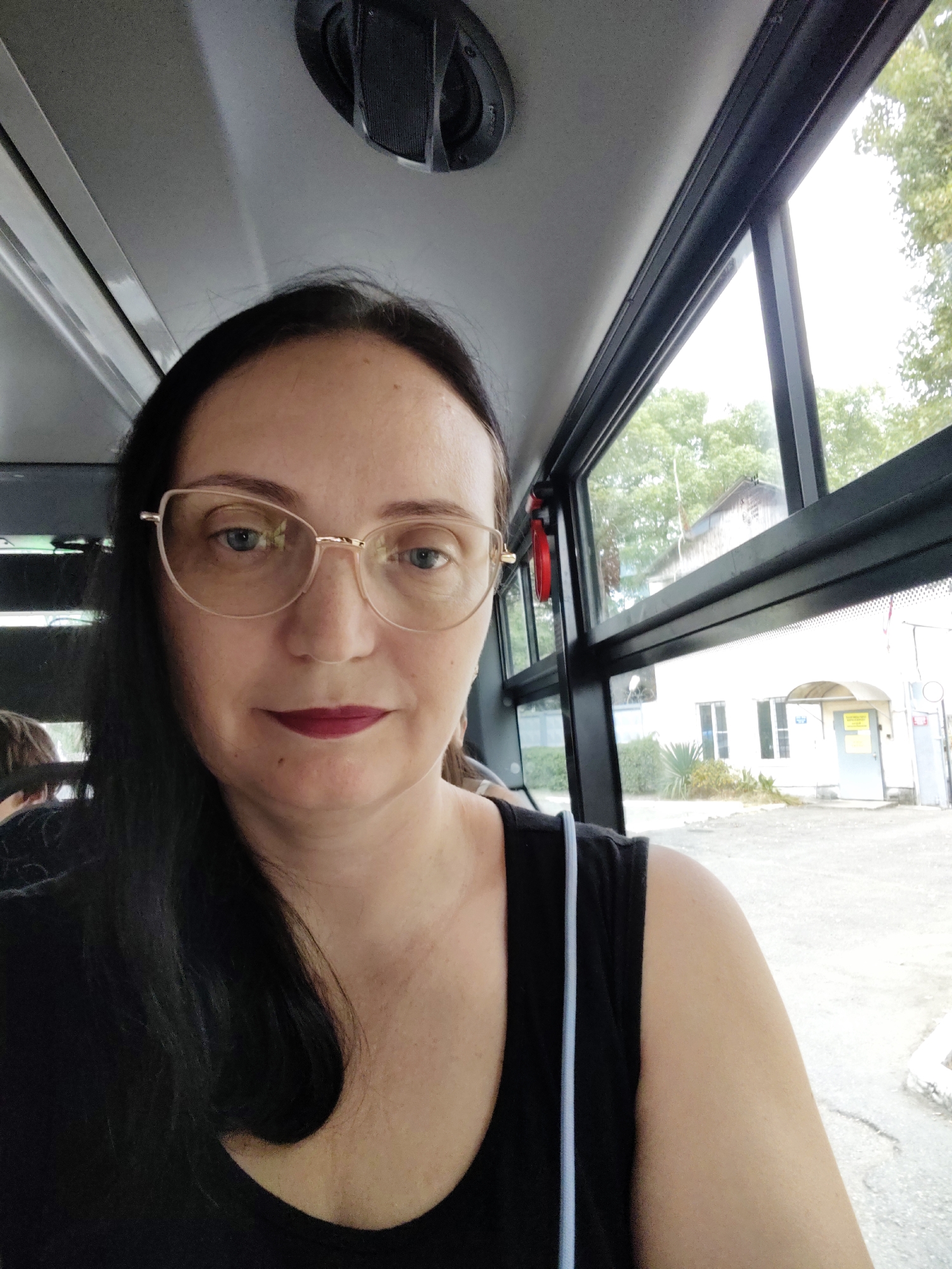 A medium shot captures a woman with dark hair and glasses inside a bus, with a view of a building and trees through the window. The woman is looking towards the camera, her expression neutral. She is wearing a black top and lipstick. The interior of the bus shows the ceiling and a speaker. Outside, a white building with a light blue door is visible, surrounded by greenery. The scene appears to be from a sunny day. The location is identified as Sochi, Russia, likely a street scene visible from the bus.