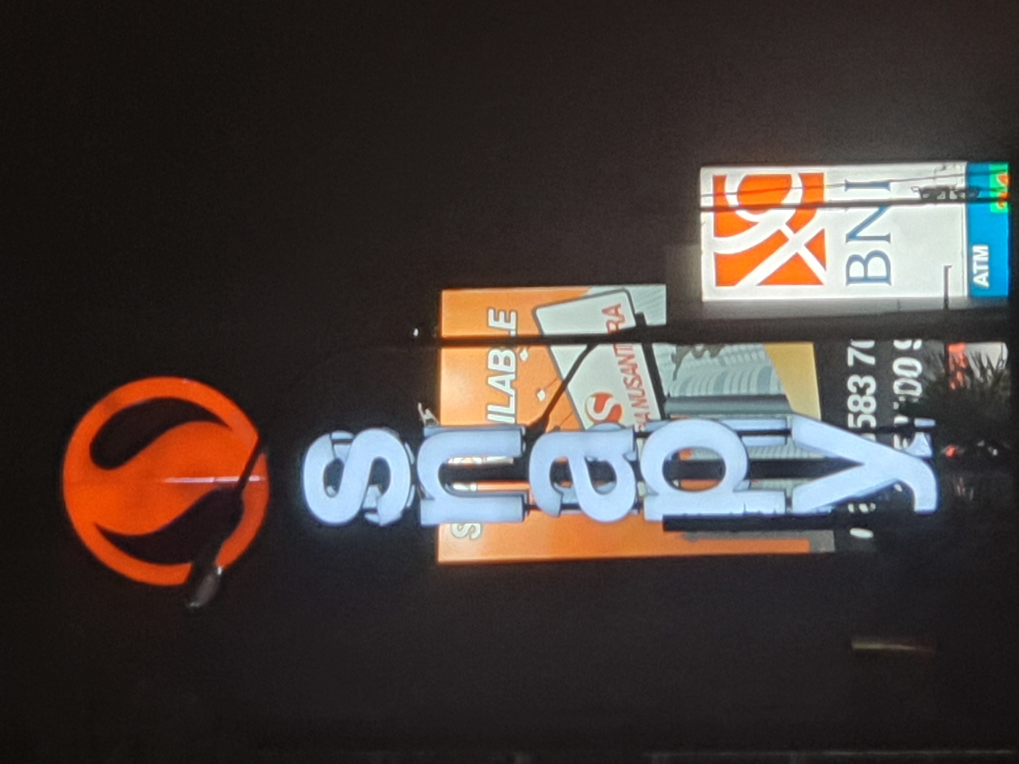 This image shows a vertical neon sign that spells out "snappy" in large white letters against a dark background. Above the word "snappy," there is a circular orange logo. To the right of the "snappy" sign, there is a rectangular orange sign with white text, partially obscured by the "snappy" sign. Below the "snappy" sign, there is a smaller sign with blue and white text that says "BNI" and "ATM". The BNI logo, which is orange and white, is visible above the text. The image appears to have been taken at night, possibly in a commercial area of North Jakarta, Indonesia, indicated by the presence of the BNI bank sign, a well-known Indonesian bank.