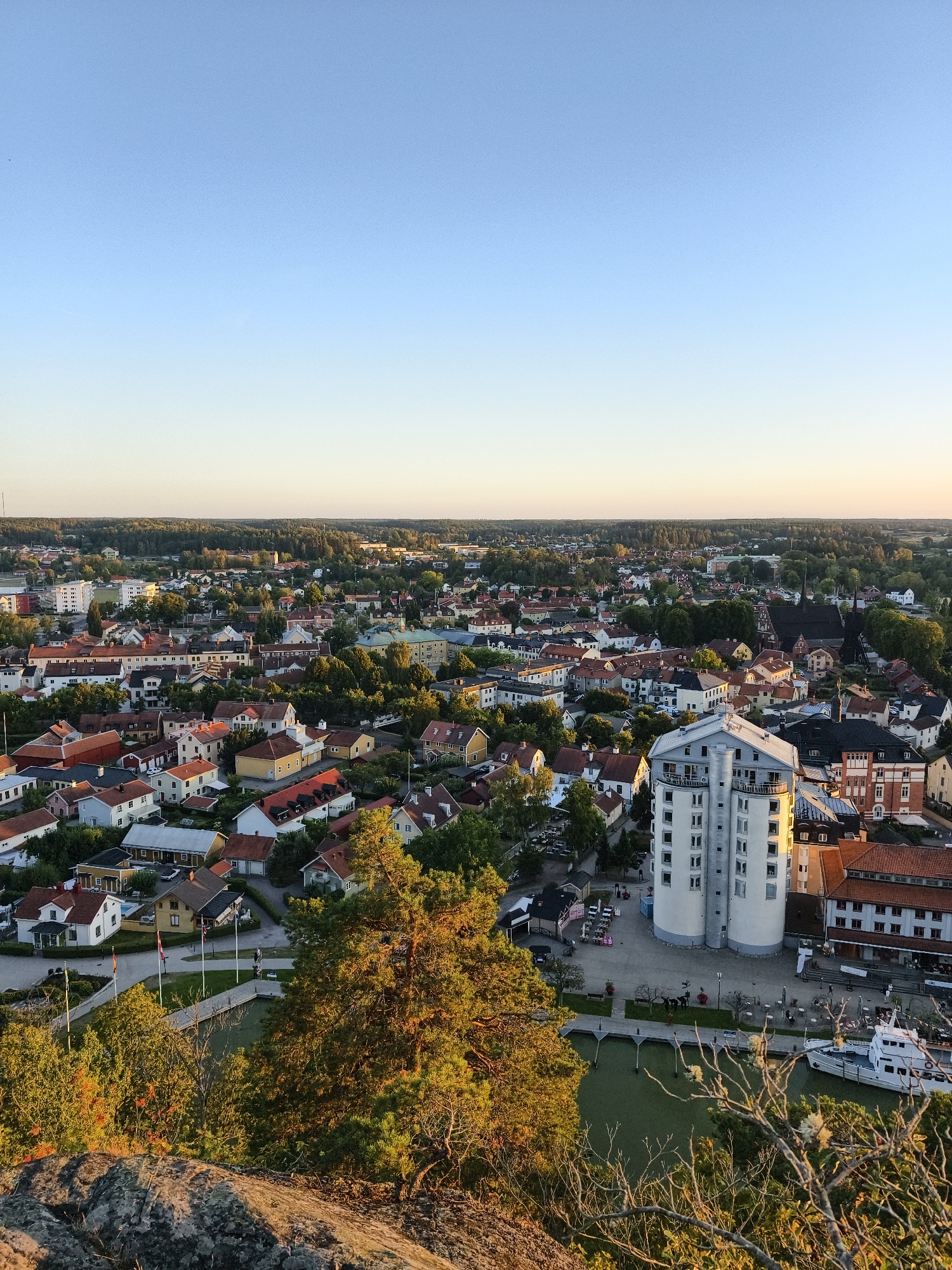 This is a high-angle, wide shot of the city of Söderköping, Sweden, taken during the golden hour of sunset. The sky is a clear, gradient blue to pale yellow, indicating late afternoon or early evening light. In the foreground, a rocky outcrop with dry, twiggy vegetation is visible, suggesting a natural viewpoint.
Below this, the city unfolds with a dense arrangement of red-tiled roofs and various architectural styles. Residential houses, many with light-colored facades, are interspersed with larger buildings and trees. A prominent, cylindrical white building with numerous windows stands out near the center-right of the image. Adjacent to it is a dock area with a white boat moored, and a body of calm water, likely a canal or harbor. Several flags are planted along a walkway leading to the dock. In the background, a dense forest borders the city, and the horizon is flat.
Scattered throughout the urban landscape are trees, some evergreen and some deciduous, with their leaves catching the warm light. There are a few distant figures visible on the walkway near the boat, and a couple of parked cars. The overall impression is of a peaceful, well-established town nestled within a natural environment.