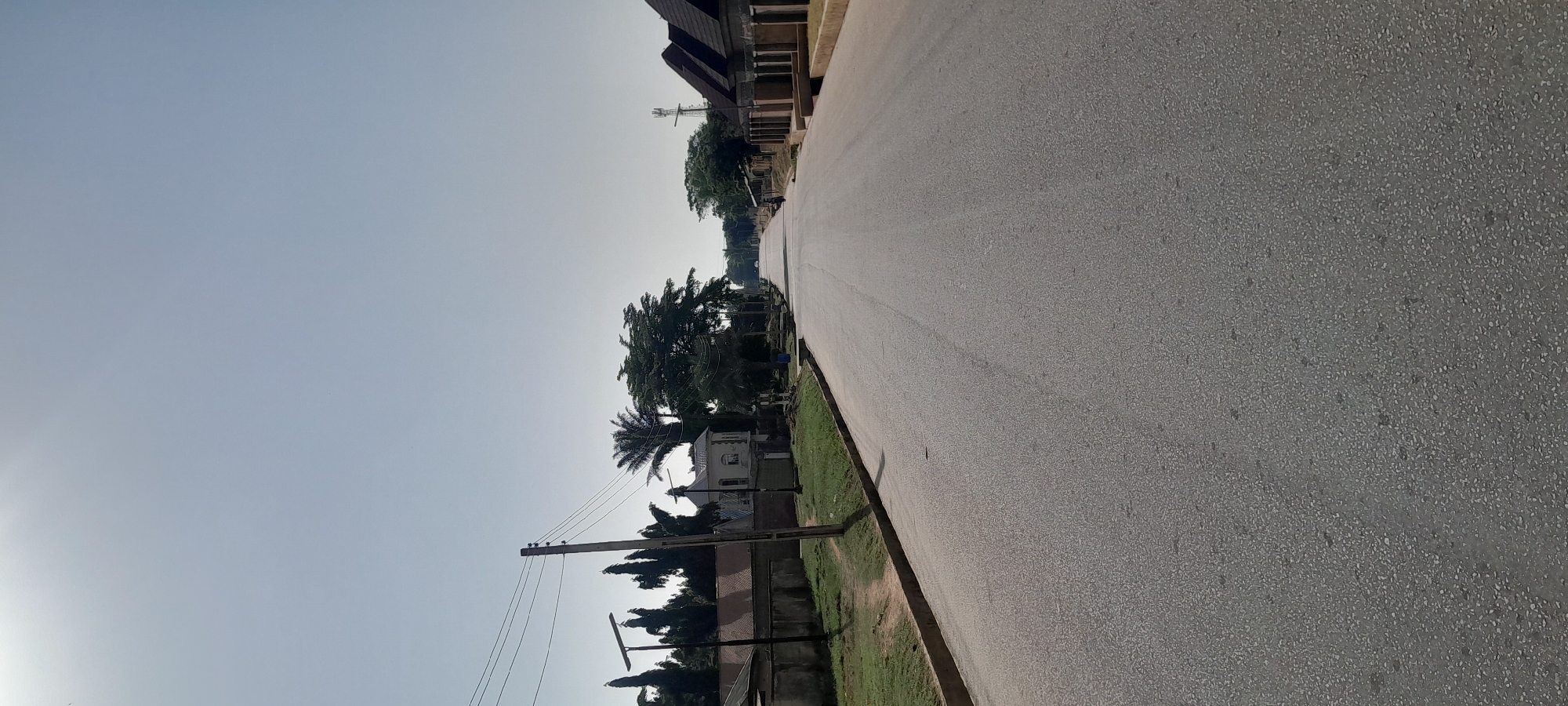 The image depicts an asphalt road in Jalingo, Nigeria, with a clear, pale blue sky overhead. The road extends into the distance, curving slightly to the right. On the left side of the road, there are houses with fences, trees, and utility poles with overhead wires. A white building with an arched entrance is visible. On the right side, there are houses with dark, sloped roofs and some are under construction. A lone motorcyclist is seen in the distance on the right side of the road. The environment appears to be a residential area with well-maintained infrastructure.