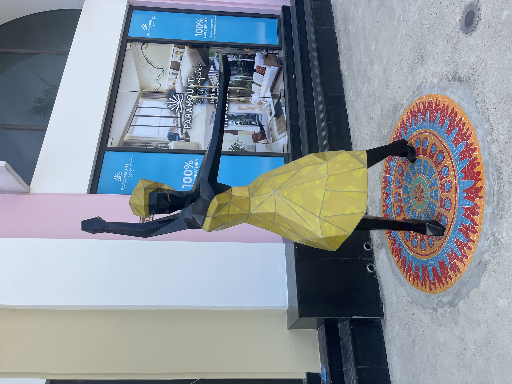 Here's a description of the image:
The image depicts a low-angle shot of an artistic installation situated outside a building. The installation is a stylized sculpture of a woman dressed in a yellow geometric dress and black accessories, positioned on a circular mosaic pattern on the concrete pavement.
The backdrop features the building, which has a pink facade, a white architectural element, and glass windows/panels with advertising for "Paramount Signature" with information in Vietnamese. The advertisement includes imagery of interior spaces and mentions "100% VIEW BIẾN" and a hotline number. The building has a set of black steps leading up to its entrance.