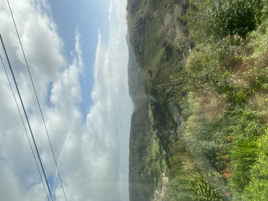 The image captures a panoramic view of a verdant, hilly landscape located in the rural vicinity of Mafra, Portugal. The upper third of the frame features a partly cloudy sky with patches of blue visible between scattered white cumulus clouds. Two dark, parallel overhead utility wires traverse the sky horizontally in the upper section. A faint, arc-shaped rainbow is discernible beneath the upper wire in the upper-left sky.
The mid-ground and background are dominated by rolling hills and valleys densely covered with dark green vegetation, including forests and cultivated fields. To the far left in a valley, a small cluster of white and light-colored buildings with reddish roofs, consistent with a village or hamlet, is visible amidst lighter green fields. A narrow, winding path or road is faintly visible traversing a hillside in the middle-right.
The immediate foreground is filled with abundant bright green foliage, including various bushes, grasses, and a cluster of red-orange flowers on the lower left. Sunlight creates several specular highlights and reflections across the foreground vegetation, indicating the presence of a reflective surface, possibly a window, through which the photograph was taken. No individuals or animate subjects are present in the scene, and no specific actions or interactions are depicted.
