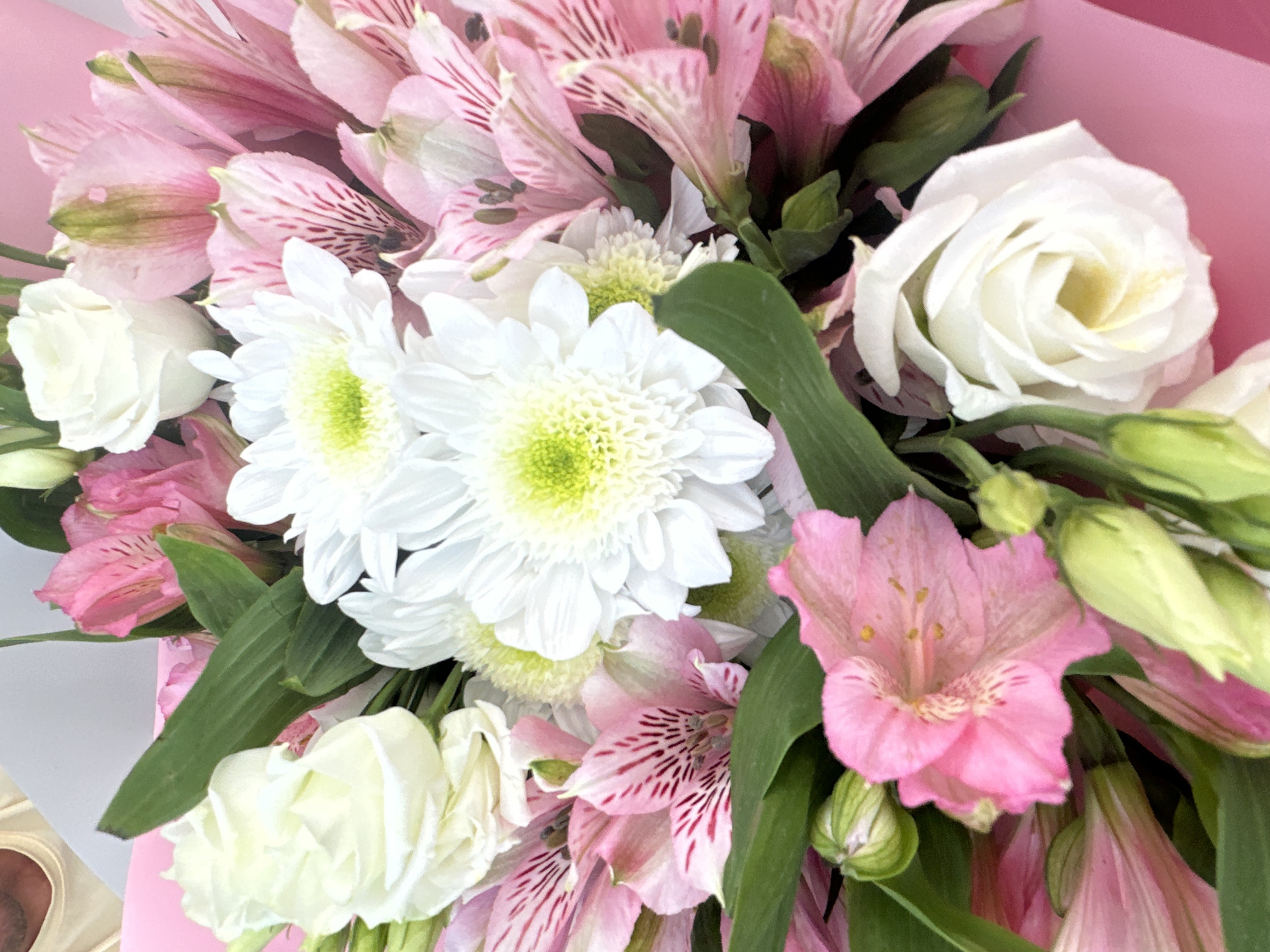 The image is a close-up, overhead shot of a vibrant bouquet of flowers, predominantly pink and white. The flowers include white roses with tightly furled petals, fluffy white chrysanthemums with lime-green centers, and delicate pink alstroemeria with darker pink streaks. Lush green leaves are interspersed throughout the arrangement. The bouquet is partially wrapped in pink paper, suggesting it's a gift or for sale. The background is a soft, light color, possibly a table or wall. There is no discernible location in Minsk, Belarus, or any people visible in the image.