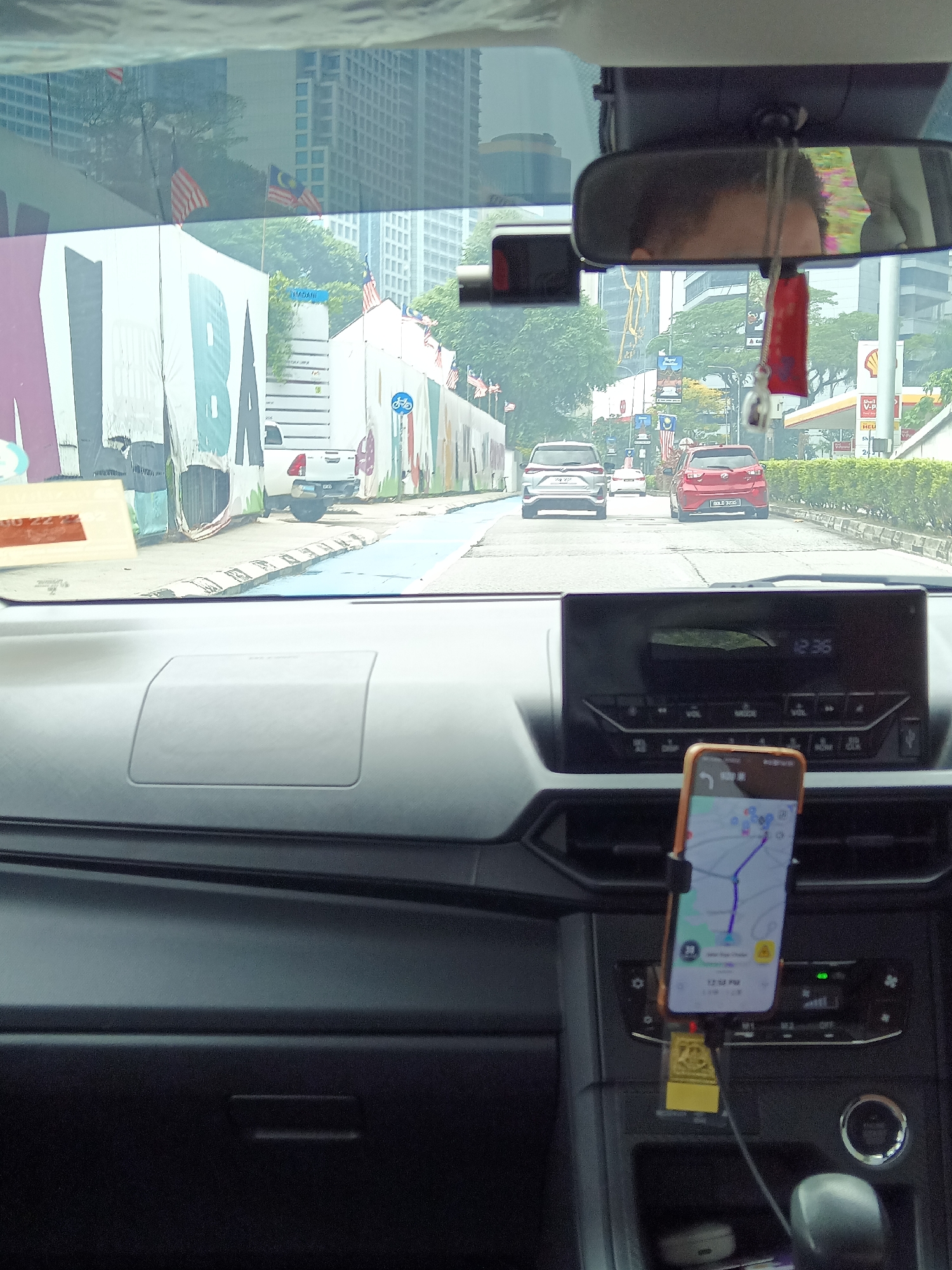 The image is taken from the interior of a car in Kuala Lumpur, Malaysia. The view is through the windshield, showing a street scene. In the foreground, the dashboard of the car is visible, with a smartphone mounted on a holder, displaying a navigation app with a map and route. The time on the phone shows 12:34 PM. A car's clock on the dashboard displays 12:36.
Outside the car, the road is lined with buildings and trees. Several cars are visible on the road, moving in the same direction. There are Malaysian flags displayed along the roadside. On the left side of the road, a large white wall with colorful graffiti is partially visible. A white pickup truck is parked behind the graffiti wall. A building with a Shell gas station canopy is visible on the right side of the road, with street signs and traffic lights. The sky is overcast. The overall impression is of a city street scene during the day, likely in a commercial or residential area of Kuala Lumpur.