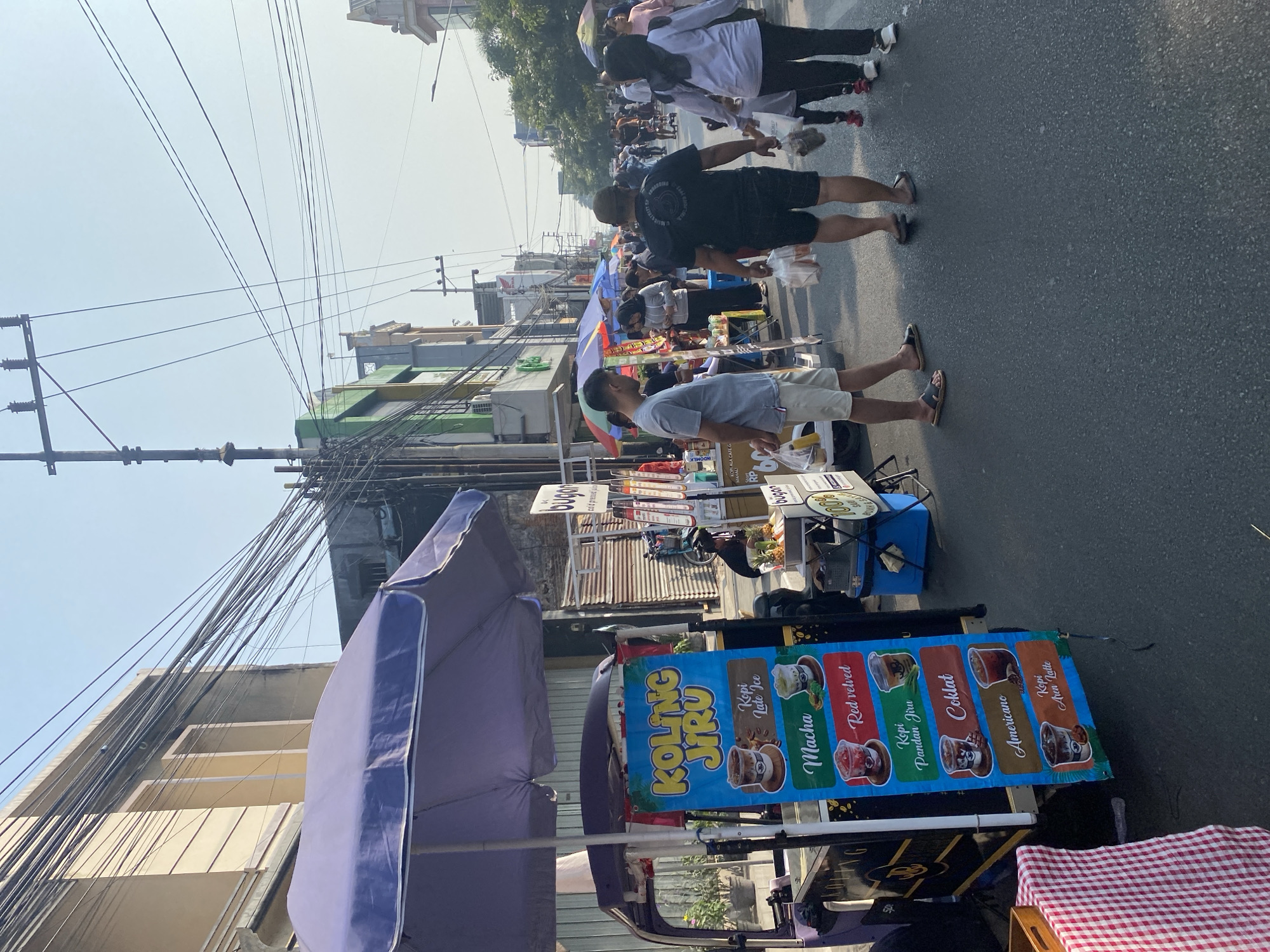 The image captures a street scene in Kediri City, Indonesia, characterized by a bustling market atmosphere. In the foreground, a small cart with a purple umbrella displays a menu advertising various coffee drinks, including "Kopi Jiru," "Macha," "Red Velvet," "Kopi Pandan Jiru," "Coklat," "Americano," and "Kopi Aren Latte." A person in a grey t-shirt and shorts stands near the cart, facing away from the camera.
The street is lined with shops and temporary stalls, with many people walking and browsing. Overhead, a dense network of electrical wires crisscrosses the sky, a common sight in urban Indonesian settings. The buildings on either side of the street appear to be a mix of older and more modern architecture. The lighting suggests it is daytime, possibly late afternoon, with the sun casting shadows. The overall impression is one of a vibrant local market, likely a popular spot for residents and visitors alike to enjoy street food and drinks.