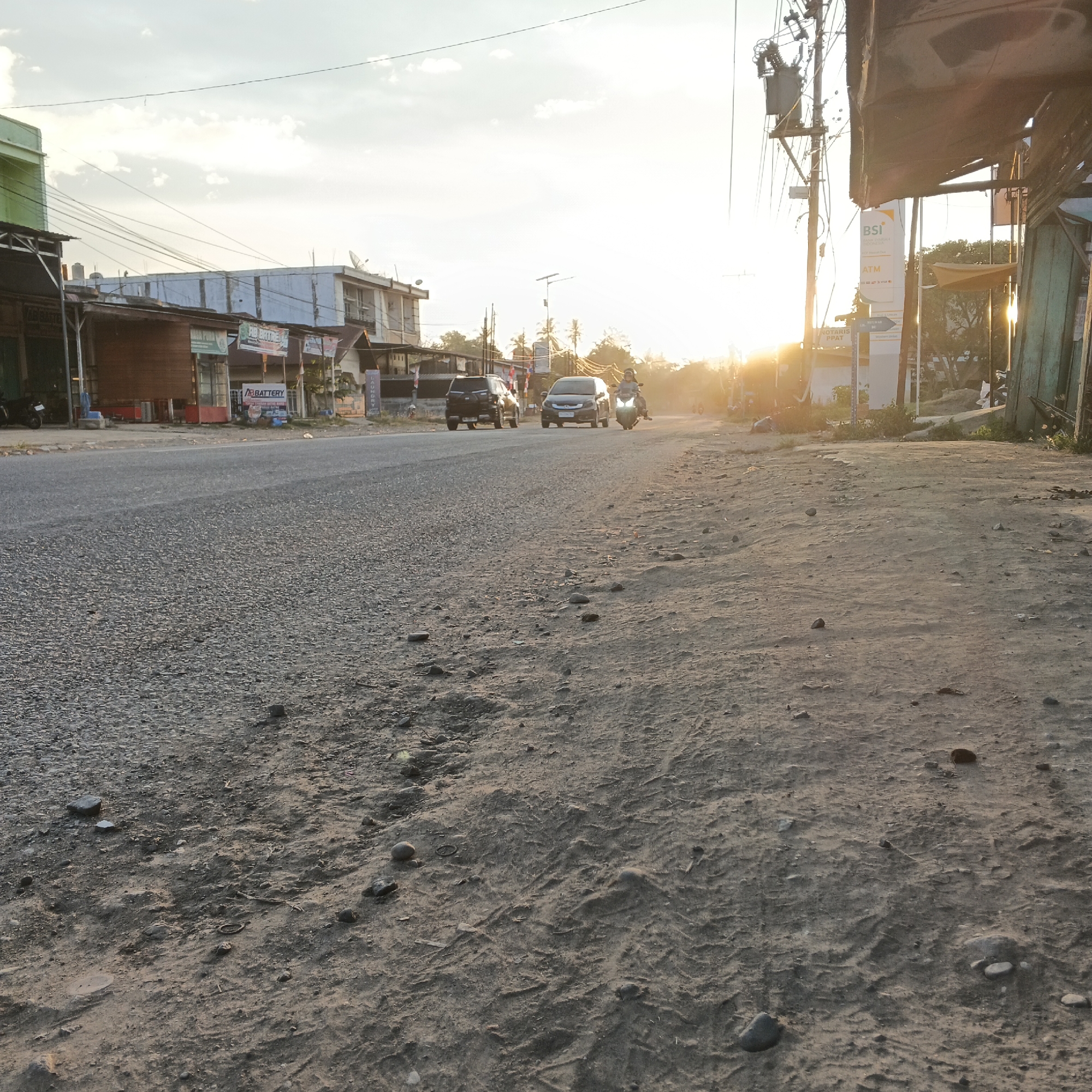 The image depicts a roadside scene in Meunasah Raya, Indonesia, during sunset. The foreground is dominated by a dirt and gravel shoulder of an asphalt road. On the road, several vehicles are visible: a black SUV, a black sedan, and a motorcycle with a rider. Buildings line the left side of the road, featuring storefronts with signage, including "AB BATTERY" and what appears to be an ATM sign for "BSI BANK SYARIAH". Utility poles with numerous wires are prominent on the right side of the image, one of which has a transformer. The sky is bright with the setting sun, casting a warm glow and lens flare across the scene, and there are scattered clouds. The overall impression is of a typical urban or semi-urban environment in Indonesia transitioning from day to evening.