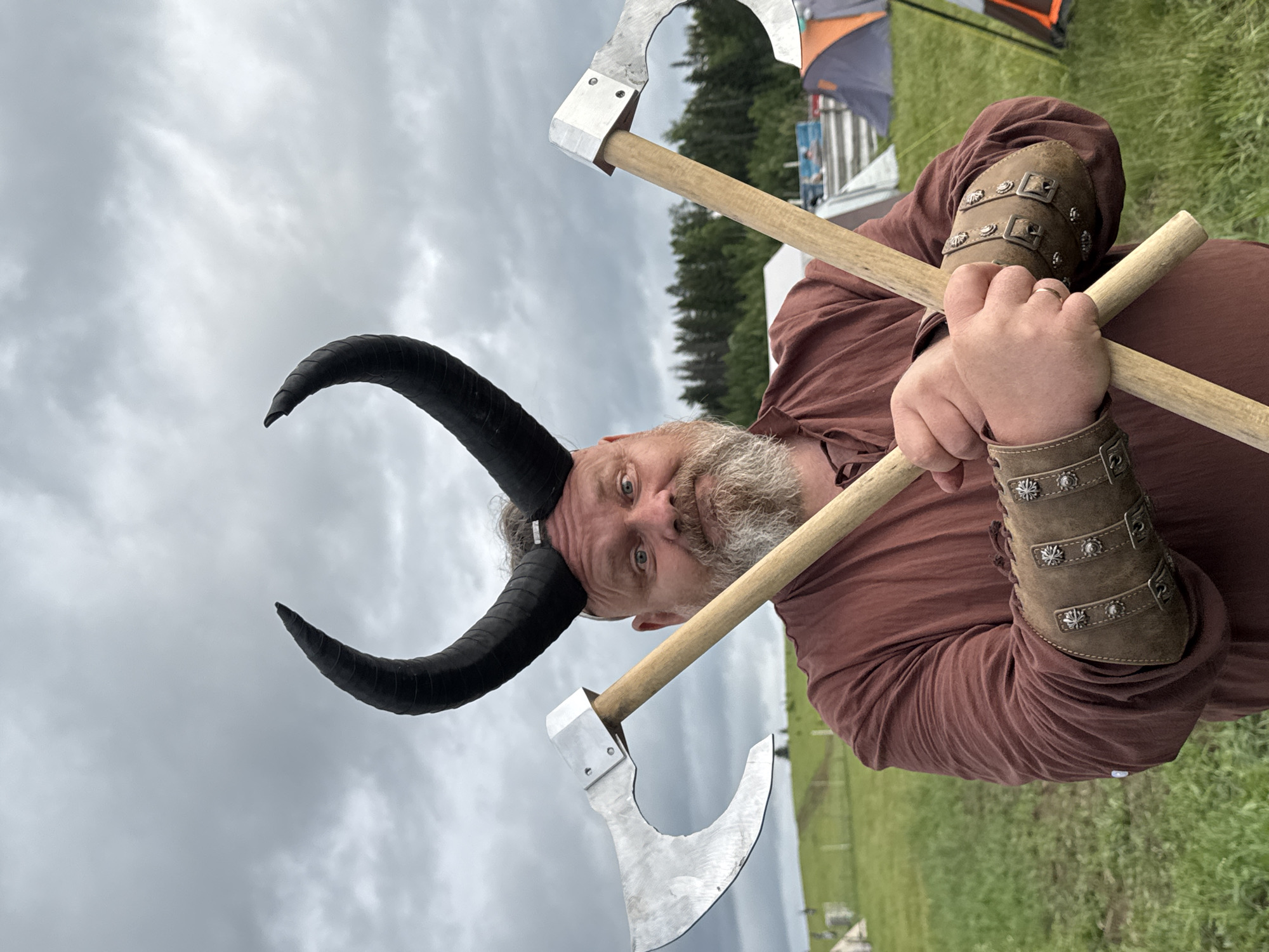 An outdoor, eye-level shot depicts a man in a costume, facing forward with a slight smirk or grin. He has a full beard and is wearing dark, curving horns on his head. His attire includes a plain brown, long-sleeved tunic and brown leather wrist cuffs (bracers) adorned with metal studs and buckles. He wears a simple ring on the ring finger of his left hand. The man is holding two axes, crossed in front of him. The axes have natural wood handles and wide, polished silver-colored metal blades.
The background shows an open grassy field with several green trees in the mid-ground. The sky is overcast with light grey clouds, indicating daytime but not direct sunlight. In the far background, partial views of what appear to be tents or temporary structures are visible, suggesting an event or festival is taking place. The overall setting is an open-air gathering, possibly a historical reenactment or fantasy-themed event, in the area of Большая Кивара, Russia. No visible text is present in the image.