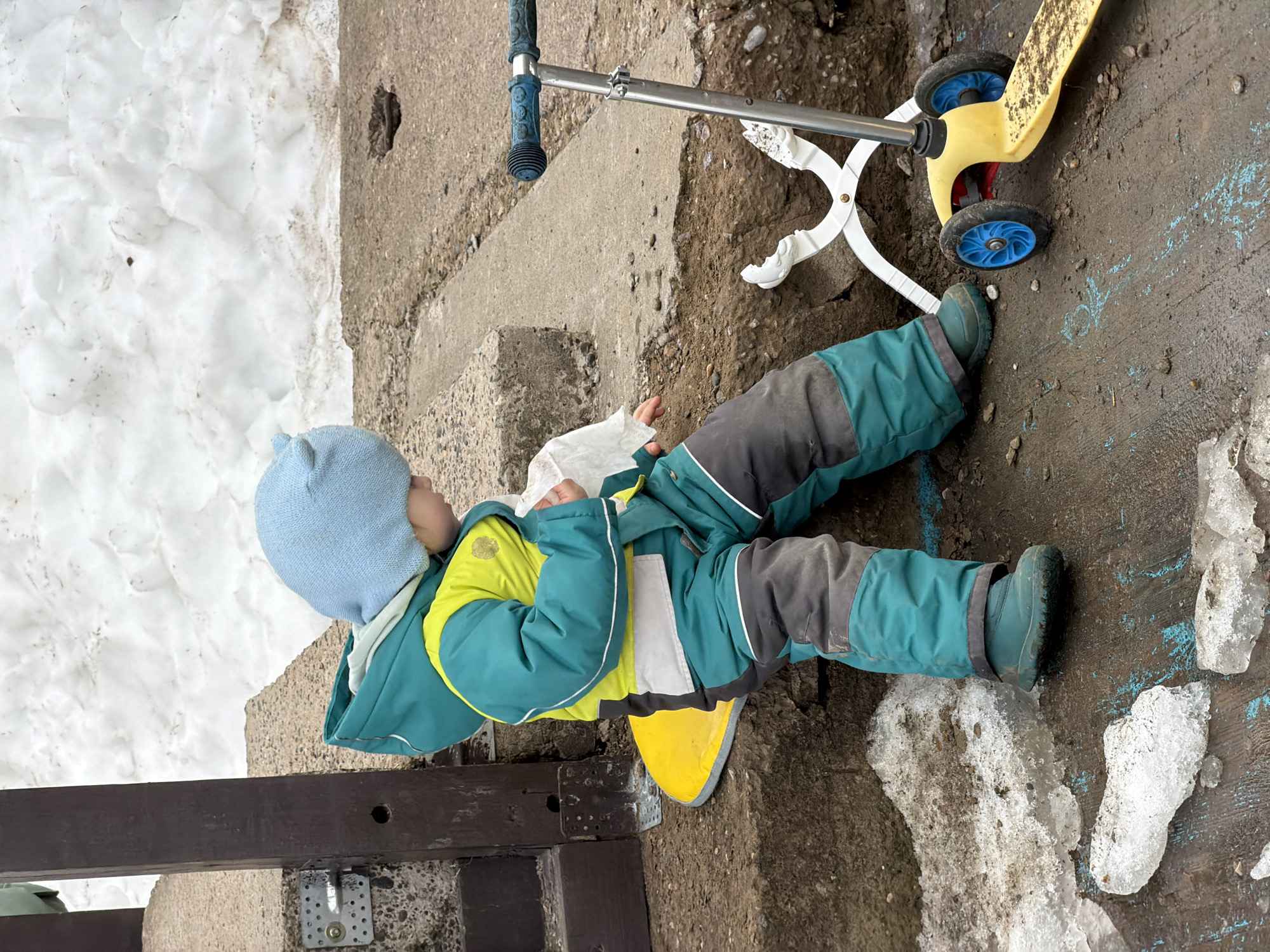 The scene depicts an outdoor environment in Chaikovsky, Russia, during a period of thaw. A young child, attired in a light blue knit hat, a two-tone teal and neon yellow hooded jacket, teal and dark grey snow pants, and teal boots, is seated on a bright yellow plastic saucer sled. The child is holding a white tissue or piece of paper in their right hand, with their gaze directed downwards and slightly to the right.
To the child's right, a yellow kick scooter with blue wheels and a blue handlebar grip rests on the muddy ground. Adjacent to the scooter, a pair of white, open snowball tongs is partially submerged in a shallow dirt trench. The ground immediately surrounding the child consists of dark brown soil, muddy patches, and scattered pieces of melting, dirty ice. Blue paint marks are visible on the wet ground in the foreground.
Behind the child, a section of cracked and worn concrete pavement is present. To the child's left, a dark brown wooden post, secured with metal brackets into a concrete base, is visible. The background features a large, irregular bank of white snow, showing signs of melting and dirt accumulation, contrasting with the exposed earth in the foreground.