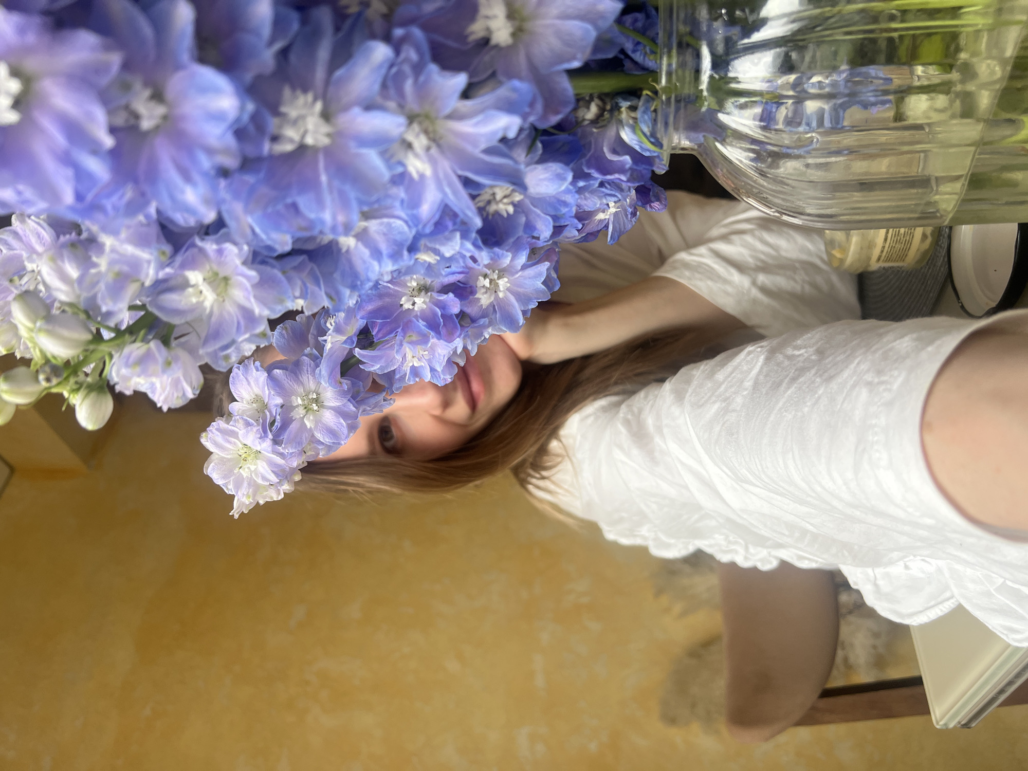 A close-up, rotated image captures a vibrant bouquet of light purple and blue flowers, likely delphiniums, and a young woman. The flowers dominate the left side of the frame, showcasing their delicate petals and white centers. They are held in a clear glass vase, partially visible at the top right, filled with water.
A woman with long, straight brown hair is partially visible on the right side of the image. Her face, featuring light-colored eyes and a soft, slight smile, is framed by her hair and partially obscured by the flowers, creating an intimate and natural pose. She is wearing a white, textured garment, possibly a loose top or dress.
The background behind the woman and a white book (partially visible in the bottom right corner) is a warm, solid yellow or ochre wall, indicating an indoor setting. The lighting is bright and natural, suggesting it is daytime. The woman appears to be looking directly at the camera, possibly taking a selfie or being photographed from an unconventional angle, with the flowers strategically placed to add visual interest. The overall impression is one of freshness, tranquility, and natural beauty. No legible text is visible in the image.