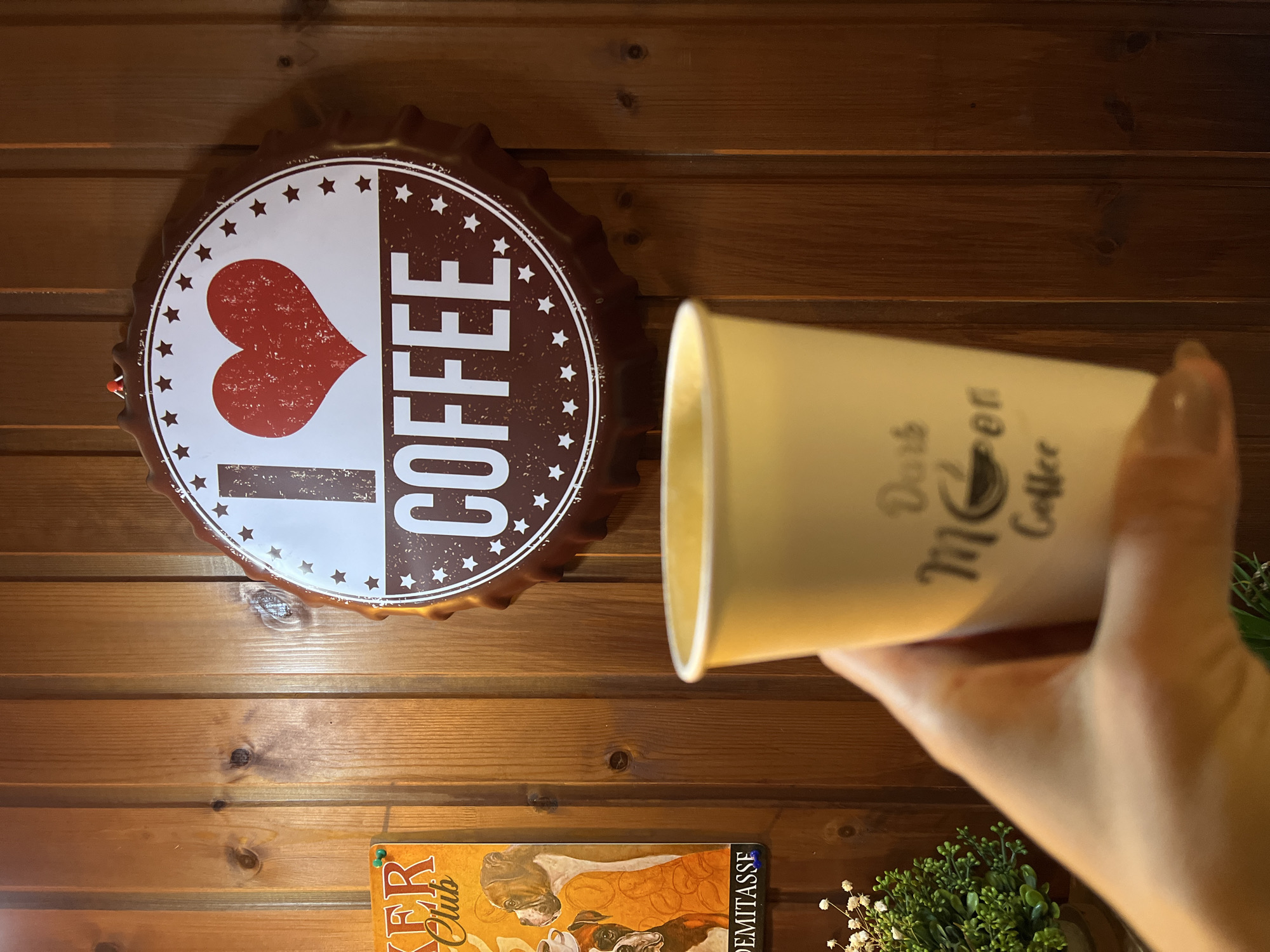 A well-lit indoor scene shows a wooden paneled wall with a decorative metal sign that resembles a bottle cap, split down the middle. The left side of the sign is white with a red heart and the right side is brown with the word "COFFEE" in white capital letters. The edge of the sign is a serrated metal that appears to be brown. A tan paper coffee cup is held by a human hand in the right side of the frame. The cup has writing on it in black ink. Below the sign, partially obscured by the coffee cup, is a poster of boxer dogs drinking coffee. Small white flowers and a small green plant are also visible. There is no information to connect the image to Petropavlovsk-Kamchatsky, Russia.