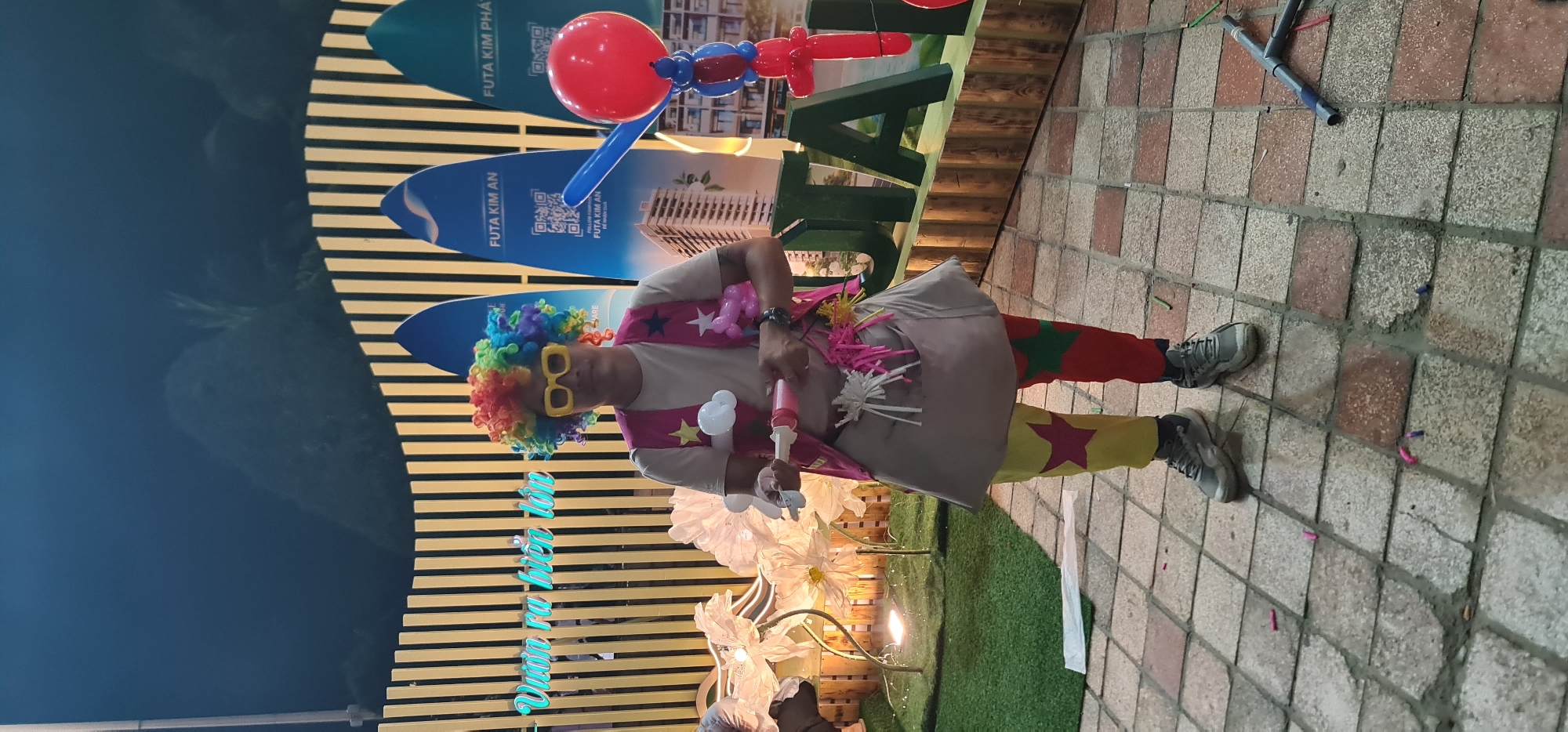 Here's a detailed description of the image:
**Scene:**
The image shows a clown performing at an event, likely an opening or promotional event, possibly for a real estate project. The event appears to be held outdoors, in a public space.
**People:**
* **Clown:** The primary subject is a clown dressed in full costume. He's wearing a colorful rainbow wig, large yellow glasses, a vest with star decorations, clown pants with red and yellow sections marked with green and red stars, and sneakers. He is holding a balloon pump and creating balloon animals.
**Objects:**
* **Balloons:** Several balloons are visible, including a red balloon, twisted balloon animals, and partially made balloons.
* **Real Estate Branding:** A backdrop shows the words "Futa Kim An" and "Futa Kim Pha" on blue surfboard shaped signs, suggesting a real estate development project. A building visual and QR code are also part of this backdrop.
* **Pavement:** The clown stands on a paved area with square tiles in shades of brown and gray.
* **Decorations:** There are floral light decorations near the clown.
* **Other:** A large green logo (partially visible) spells out “Futa Kim An” and is decorated with a balloon sculpture.
**Environment:**
The background appears to be a mix of constructed elements, including wall of vertical gold and black stripes. The setting is likely outdoors, in a public space.
**General Impression:**
The scene conveys a festive atmosphere associated with a grand opening or promotional event. The clown is the main attraction, engaging with potential attendees through balloon art. The branding strongly suggests a real estate context.