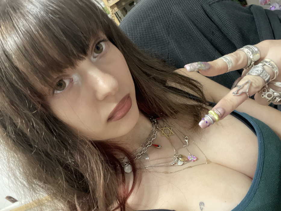 A young woman with dark brown hair and bangs poses for a selfie. She wears multiple silver rings and layered necklaces, including one with a Star of David. Her nails are long and decorated. She wears a dark green top. Her face is pale with brown eyes and light-colored makeup. A small heart tattoo is visible on her chest. The background appears to be a dark-colored couch.