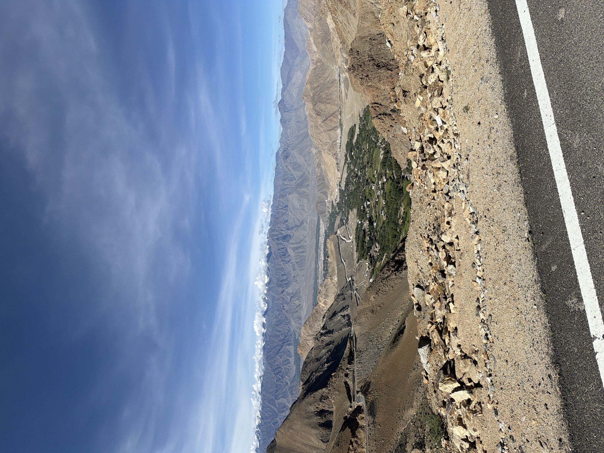 The image captures a panoramic view of the landscape surrounding Leh, India, likely from a high vantage point along a mountain road. In the foreground, a paved road with a white line runs horizontally across the bottom of the frame. Adjacent to the road, a rocky, gravelly terrain is visible, transitioning into the slopes of barren, arid mountains. These mountains exhibit shades of brown and tan, with visible geological strata.
Further in the distance, a winding road snakes through the valley. Nestled within this valley is a lush, green settlement, characterized by scattered houses and dense vegetation. This verdant area stands in stark contrast to the surrounding arid mountains.
The middle ground is dominated by a series of rugged, brown mountain ranges. These mountains appear dry and sparsely vegetated. In the background, a majestic range of snow-capped mountains forms a dramatic backdrop against a clear blue sky with scattered white clouds. The overall impression is one of vast, rugged natural beauty, with human settlements appearing as small oases within the expansive mountainous terrain.