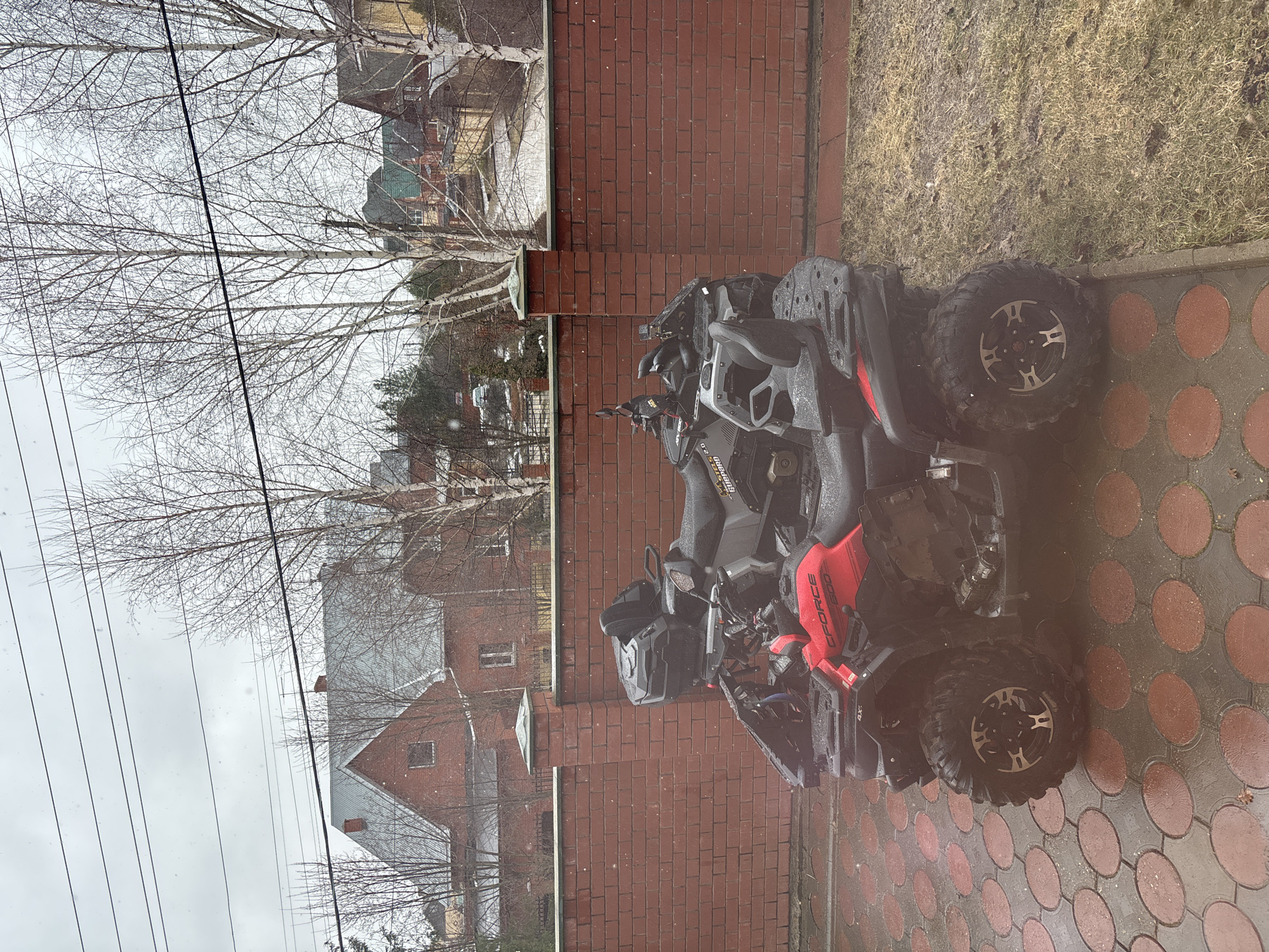 The image depicts a muddy black and red CFMOTO CFORCE 600 ATV parked on a wet brick and circular patterned surface. The ATV is positioned next to a red brick wall, with a view beyond the wall showing bare trees, residential houses, and overcast sky. The setting appears to be in a residential or suburban area. The image's details align with the city of Ferma, Russia, based on the architecture and environmental conditions. There is no evidence of people in the frame. The ATV is stationary, with no visible actions or interactions occurring.
