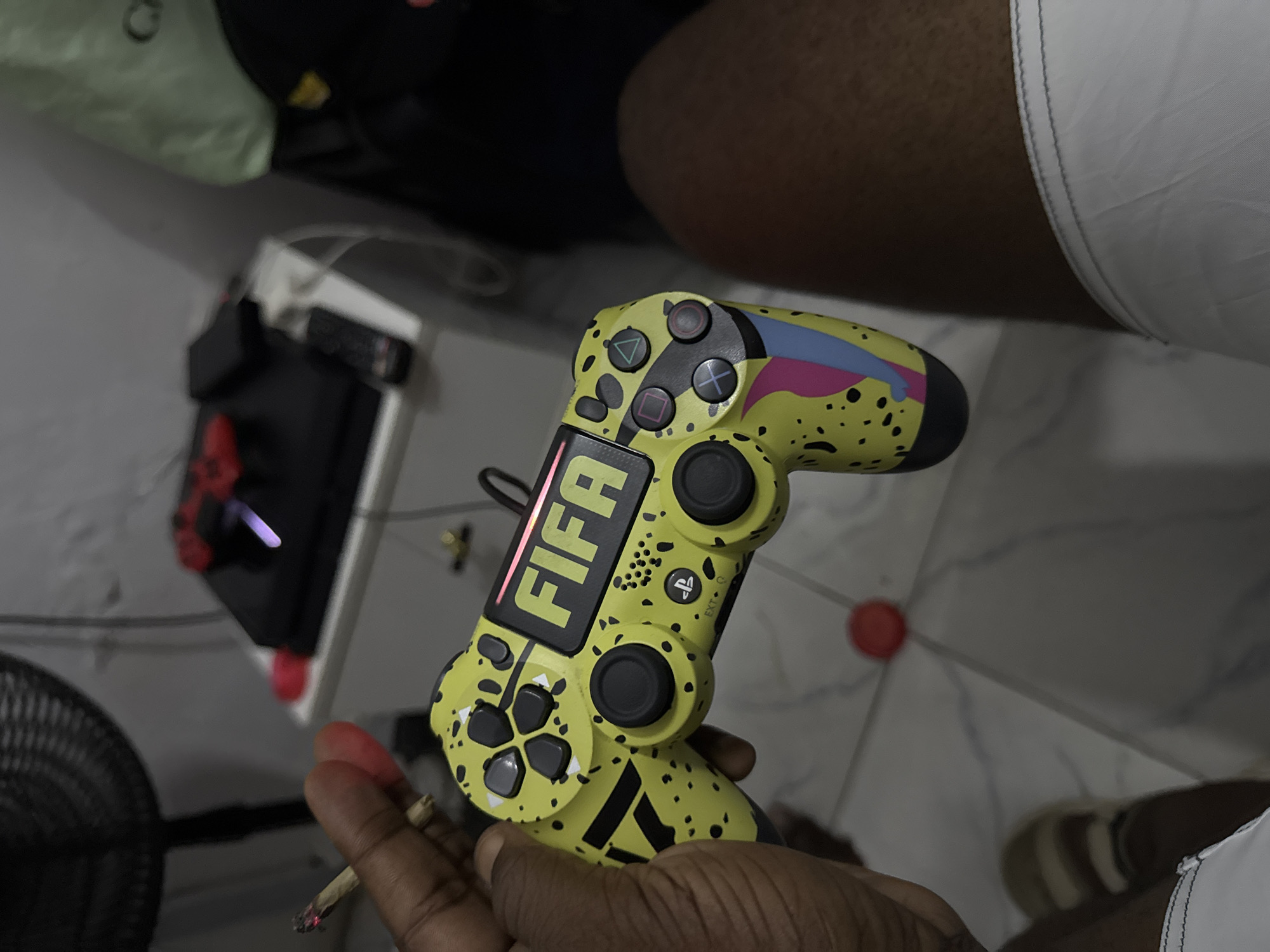 A top-down shot depicts a yellow PlayStation 4 controller with black accents, custom-decorated with a "FIFA" logo. The controller is held in a dark-skinned hand with fingers showing signs of being stained. A partially-smoked cigarette is held between the fingers. Background details include a gaming setup with a black keyboard and mouse on a white surface, and a fan. The setting appears to be indoors, likely a personal gaming space. The location is Ado, Nigeria.