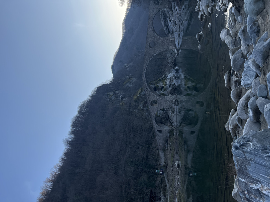 The image presents a panoramic view of Dyavolski Most (Devil's Bridge) over a river in Diadovtsi, Bulgaria. The central subject is a multi-arch stone bridge, constructed from rough-hewn grey stones, spanning the waterway. Its arches are clearly reflected in the calm, dark green water below. The riverbed in the immediate foreground features numerous grey, smooth boulders and rocks. On the left bank, adjacent to the bridge, a small open-sided wooden structure with a reddish roof is present, alongside a slender, light-colored vertical object featuring red elements, possibly a sign or marker. A steep, densely forested mountainside, predominantly with bare deciduous trees, rises directly behind the left side of the bridge and extends upward to the left of the frame. A more distant, tree-covered hill is visible to the right. The sky overhead is clear and pale blue, indicating either early morning or late afternoon light. No human figures are present, nor are any active interactions or specific actions depicted.