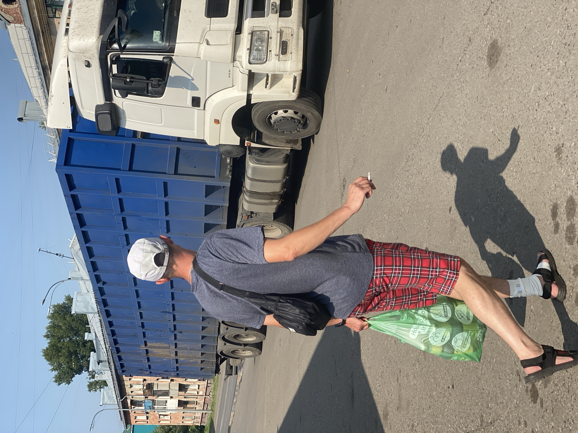 In Novokuznetsk, Russia, a person walks on a paved street next to a large blue truck with a trailer. The person, wearing a grey t-shirt, red plaid shorts, a white baseball cap, and black sandals, carries a green plastic bag filled with items and a lit cigarette in their right hand. A black sling bag is worn across their body. The truck is white with a blue container. Buildings and trees are visible in the background. The sun casts shadows on the street.