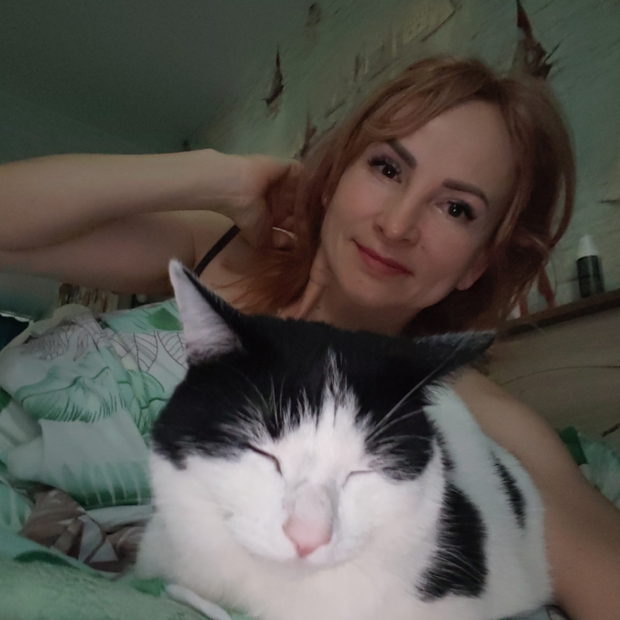 A woman with red hair is holding a black and white cat. The woman is smiling. The cat has its eyes closed. The background shows a bed with green and white patterned sheets and a wall. A bottle is also on the wall. The location is Chaikovsky, Russia.