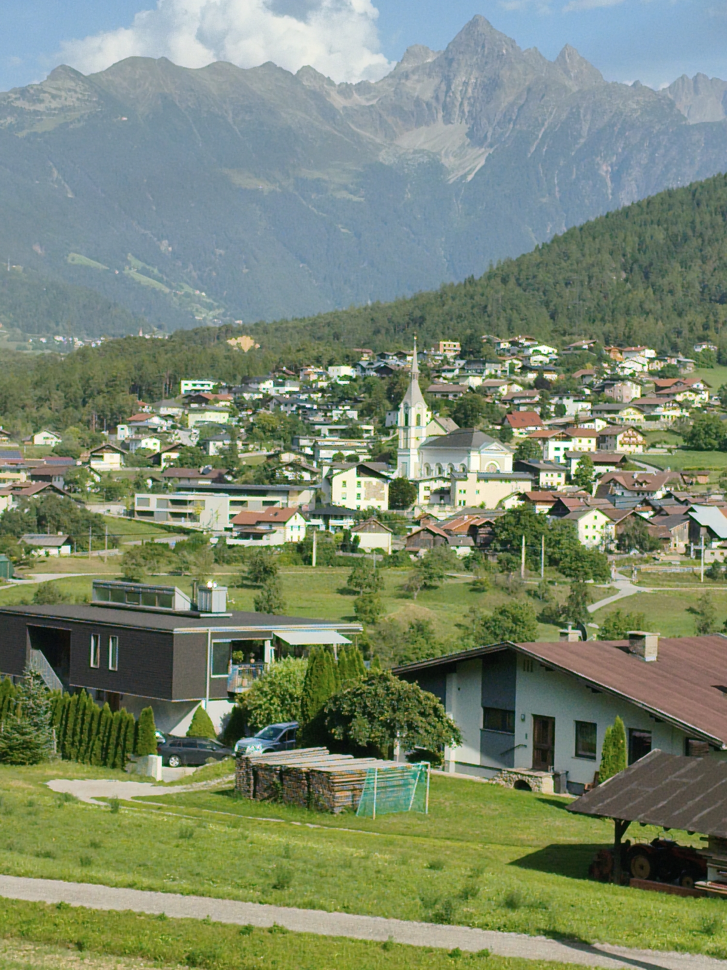 The image depicts a panoramic view of the village of Roppen, Austria, nestled at the foot of a majestic mountain range. The foreground features a grassy area with a paved path winding through it. To the left, a modern, dark-colored house with large rectangular windows is visible, with a row of tall, dark green evergreen trees bordering it. Two cars are parked on a driveway near the house, a dark sedan and a lighter colored SUV. To the right of the house, a stack of lumber and a soccer goal with a green net are situated on the grass. In the mid-ground, the village of Roppen unfolds, characterized by a collection of residential buildings with red-tiled roofs, interspersed with trees. A prominent white church with a tall steeple stands out in the center of the village, indicating its likely location in the heart of Roppen. Further back, the slopes of a lush green mountain rise steeply, dotted with more houses and trees. The background is dominated by a towering, rugged mountain range with rocky peaks, partly obscured by haze and clouds. The sky above is a clear blue with some scattered white clouds. The overall scene suggests a tranquil, picturesque alpine setting.