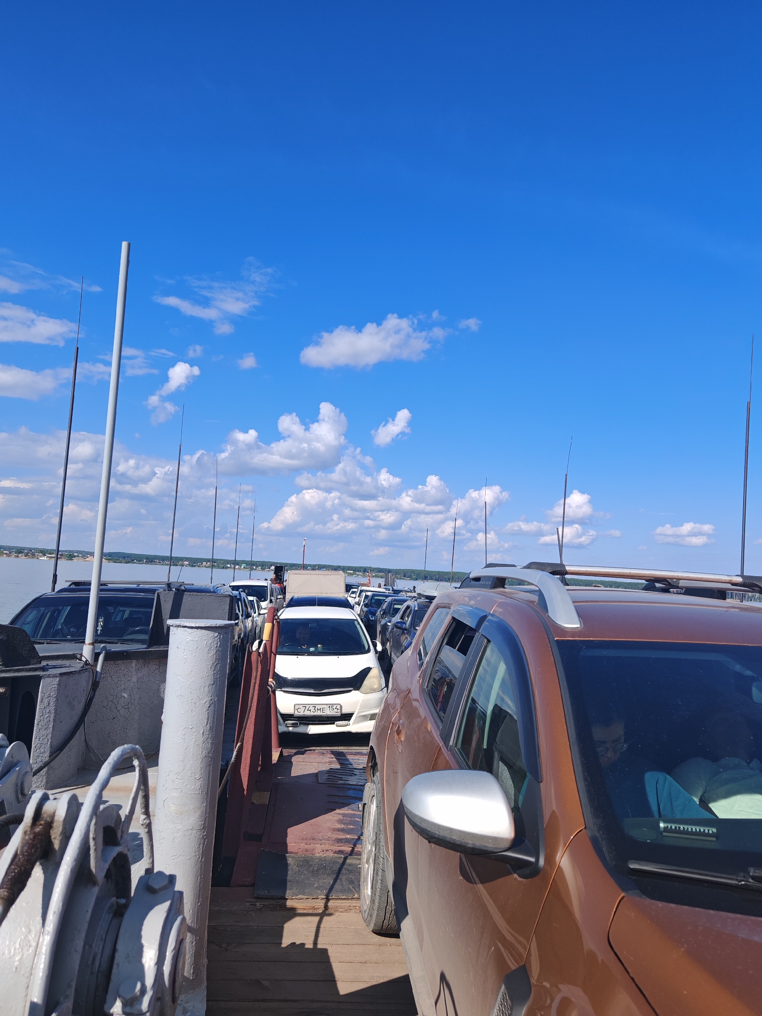 The image shows a ferry crossing a body of water, likely the river Ob, near the village of Nizhnekamenskoye, Russia. The ferry is loaded with several vehicles, including a brown Duster SUV in the foreground, a white hatchback, and other cars. A person wearing glasses is visible inside the brown SUV. The sky is blue with scattered white clouds. The ferry appears to be in motion or preparing to move, with antennas or poles visible along its length. The scene suggests a common mode of transportation for vehicles in the region.