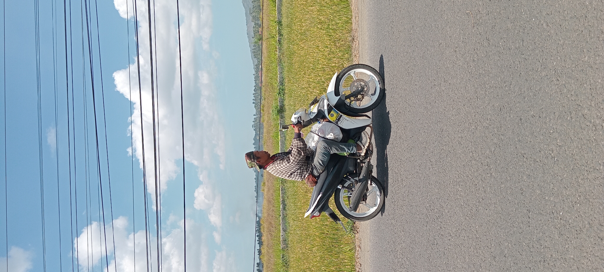 A man wearing a checkered shirt and a camouflage hat rides a white motorcycle down a paved road. The motorcycle has a black seat and a white body with black and yellow accents. The man is carrying a large white plastic bag on the back of the motorcycle, and another smaller brown item is strapped to the back of his waist. The road is bordered by a field of tall, yellow grass, likely rice paddies, and a line of trees can be seen in the distance. The sky is blue with scattered white clouds, and several power lines stretch across the upper portion of the image. The scene appears to be in Karangpawitan, Indonesia, based on the landscape characteristic of rural areas in the region.
