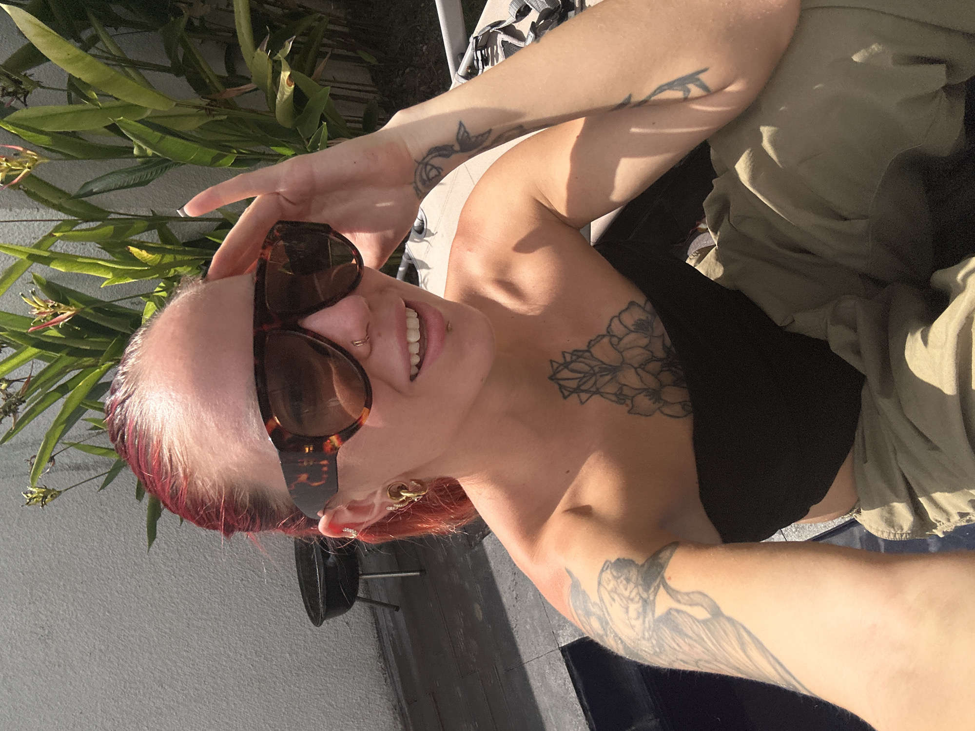 A fair-skinned woman with red hair styled back, wearing large tortoise-shell sunglasses, a black strapless top, and tan pants, is photographed outdoors. She has tattoos on her arms and chest and is wearing hoop earrings and a nose ring. Her right hand is raised to her forehead. In the background, a grey textured wall is visible, along with lush green plants. To the left, a black charcoal grill with a lid sits on a patio. The scene appears to be a backyard or patio area.