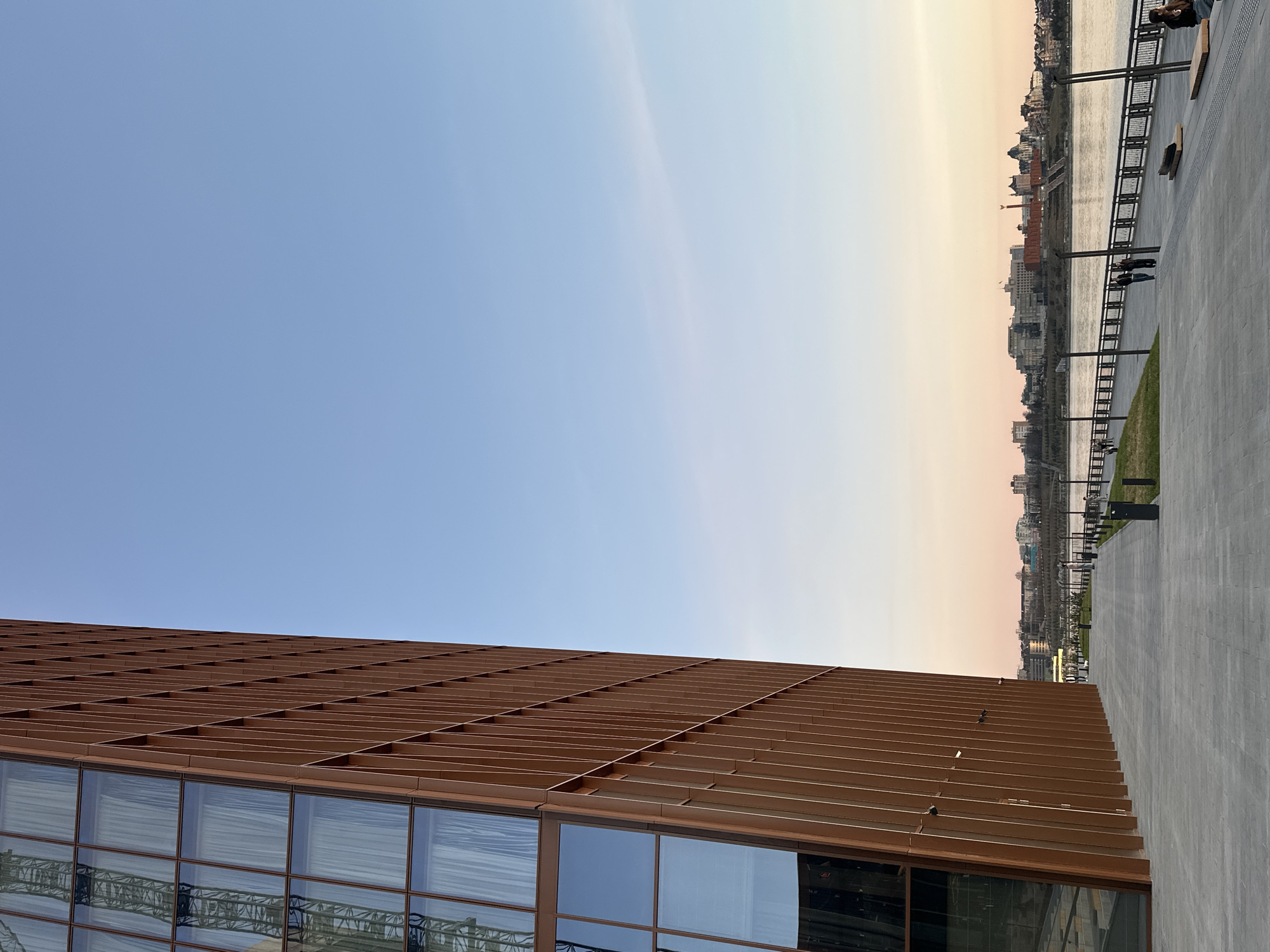This image, when viewed upright, presents a tranquil urban waterfront scene, likely during either sunrise or sunset in Kazan, Russia, given the soft, gradient sky. The upper part of the sky transitions from a clear blue directly overhead to a warm, peachy-orange glow closer to the horizon, indicating the time of day.
A prominent, modern building occupies the left side and foreground of the image. Its upper facade is distinctive, composed of numerous reddish-brown, horizontally arranged panels or slats that create a textured, grid-like pattern. Below this, large rectangular windows reflect the sky and reveal glimpses of interior structures. Several small birds, possibly pigeons, are perched on the building's facade.
To the right of the building, a wide, light-grey paved promenade or embankment stretches along the edge of a broad river. A dark metal railing with regularly spaced posts and several streetlights lines the waterfront. Across the river, a distant city skyline is visible under the soft light, featuring various buildings and structures. A few people can be seen on the embankment: one person is seated on a bench on the far right, and two other figures are walking or standing further along the promenade.
The scene evokes a calm and peaceful atmosphere of urban life by the water. There is no visible text in the media file.