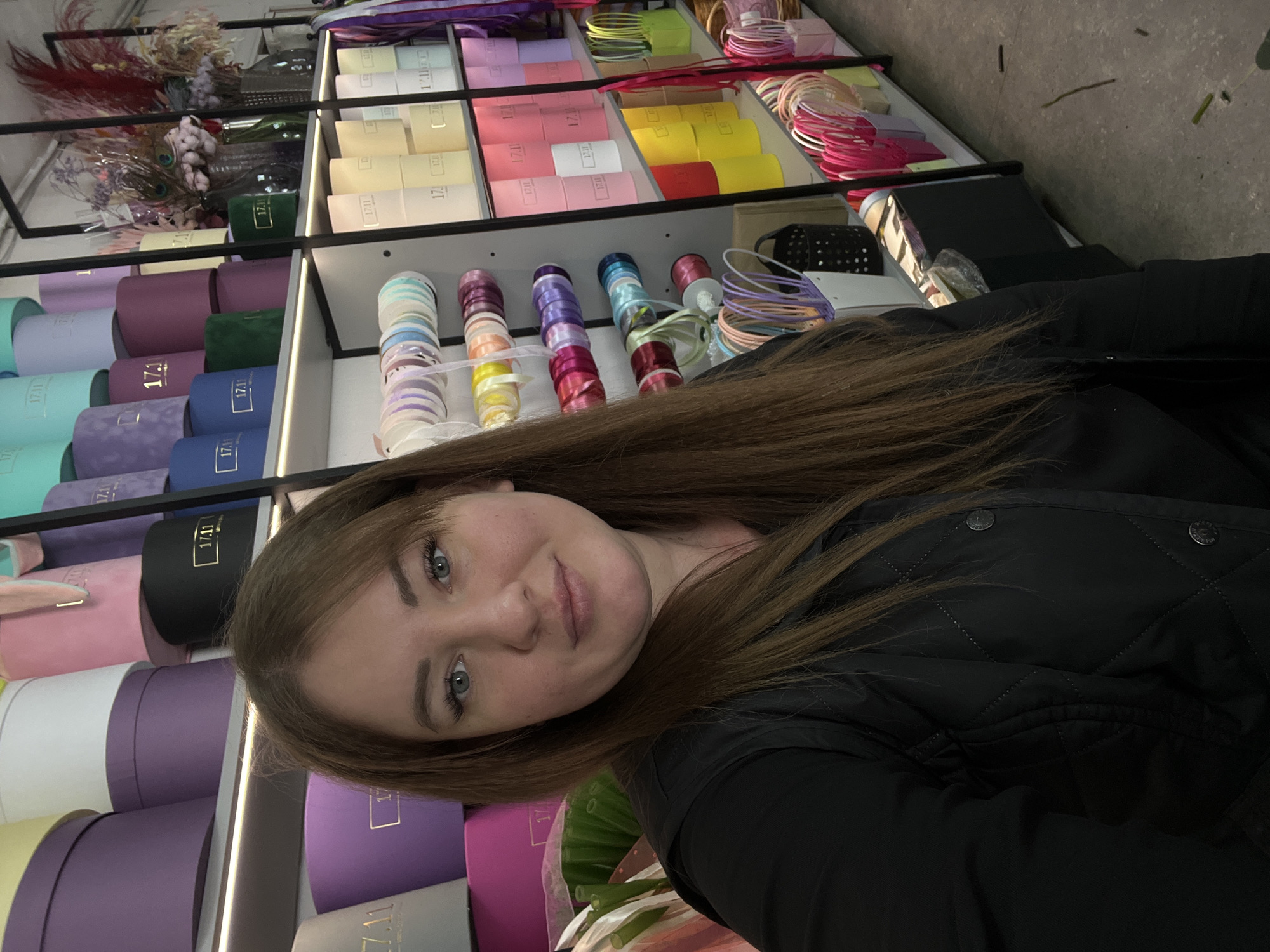 The image depicts a woman taking a selfie in what appears to be a florist shop. The shelves behind her are filled with various floral supplies, including round boxes in a range of colors, rolls of ribbon, and dried flowers. The boxes appear to be marked with the text "17.11". The woman is wearing a black jacket. The location is identified as Chaikovsky, Russia.